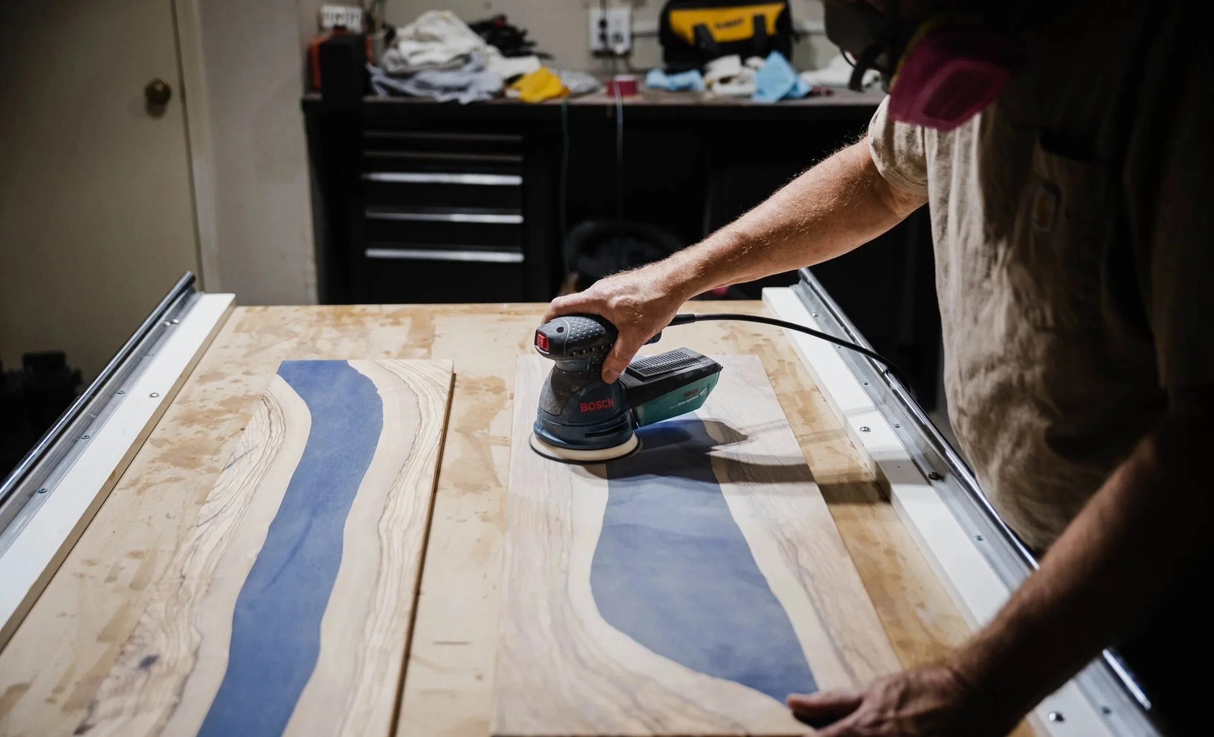 Person polishing a wooden board with a Bosch random orbit sander in a workshop.