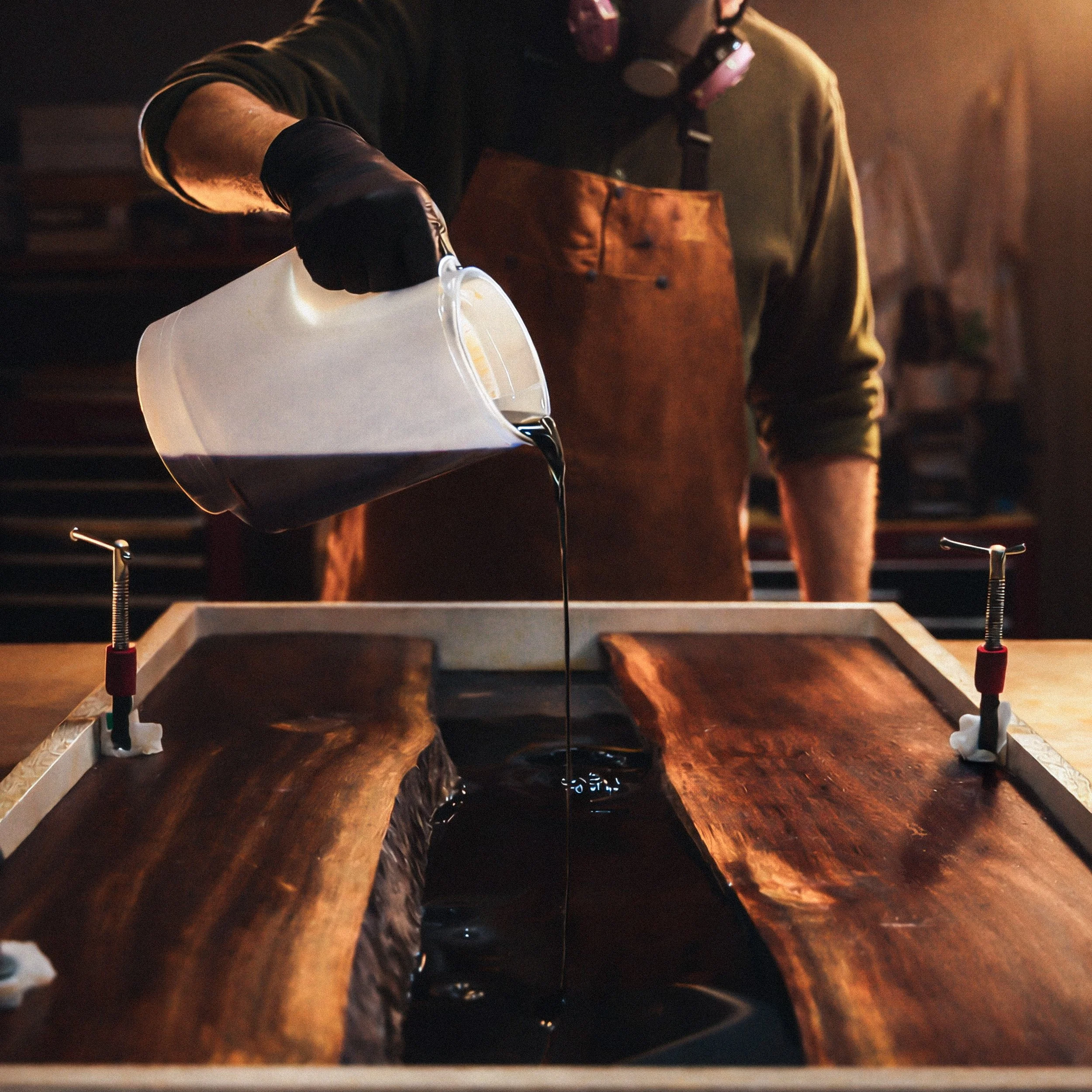 A person wearing gloves and an apron pouring resin into a wooden mold for woodworking or crafting.