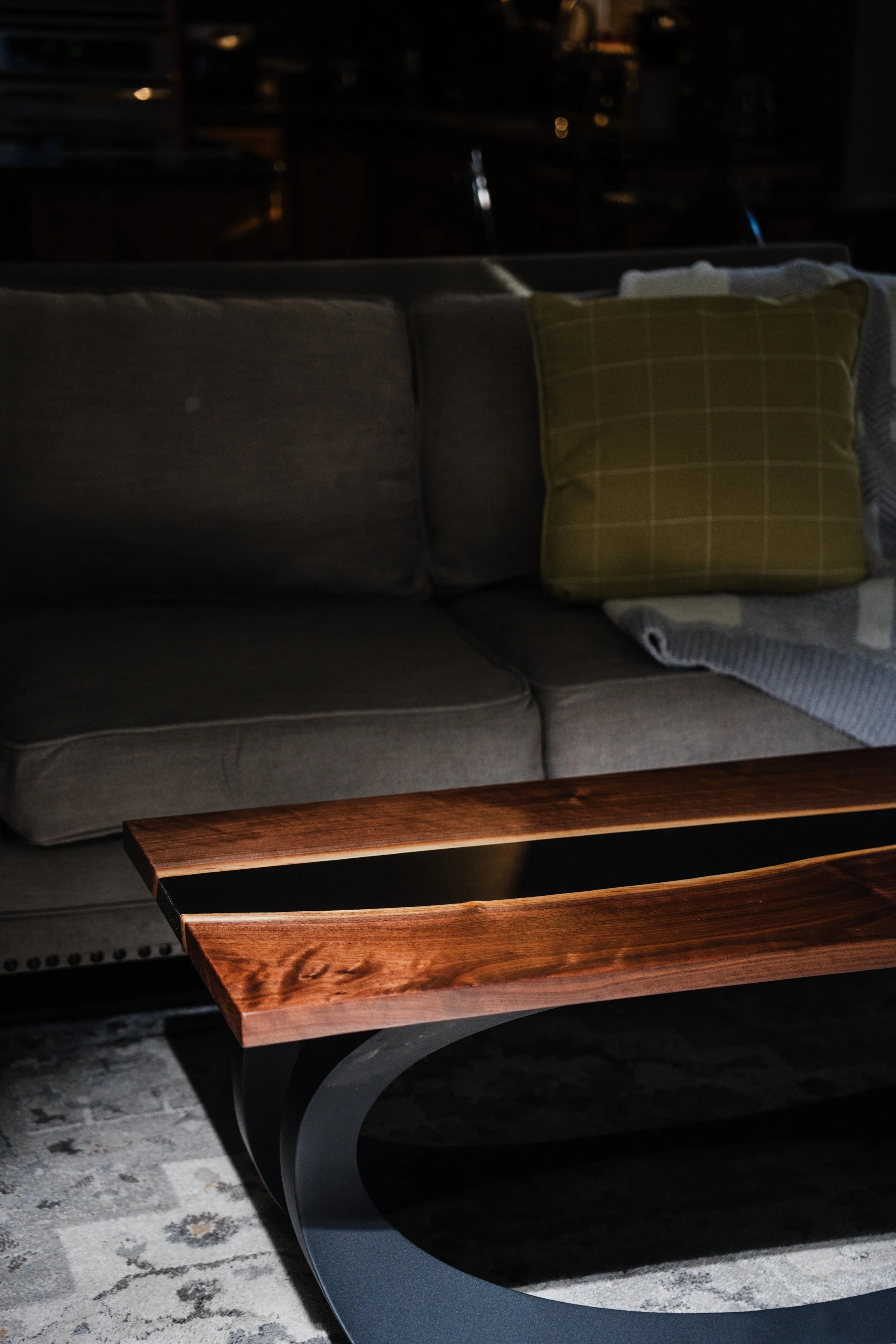 A living room scene with a dark gray sofa, a mustard yellow plaid pillow, a blanket, a wooden coffee table with a curved metal base, and a patterned rug, with dim lighting.