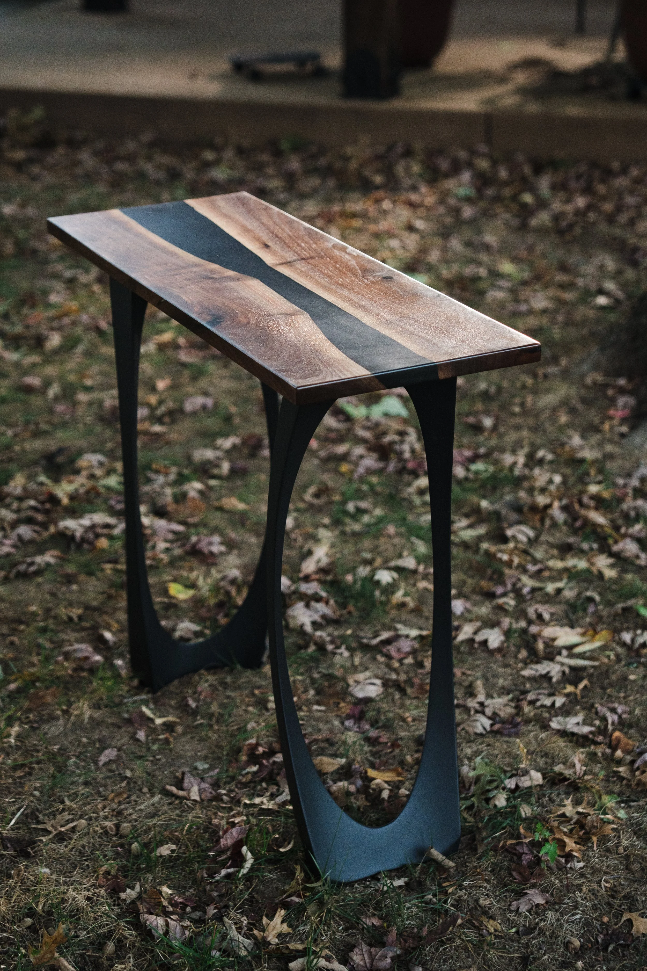 Wooden table with a black stripe down the middle standing on a patch of grass and fallen autumn leaves.