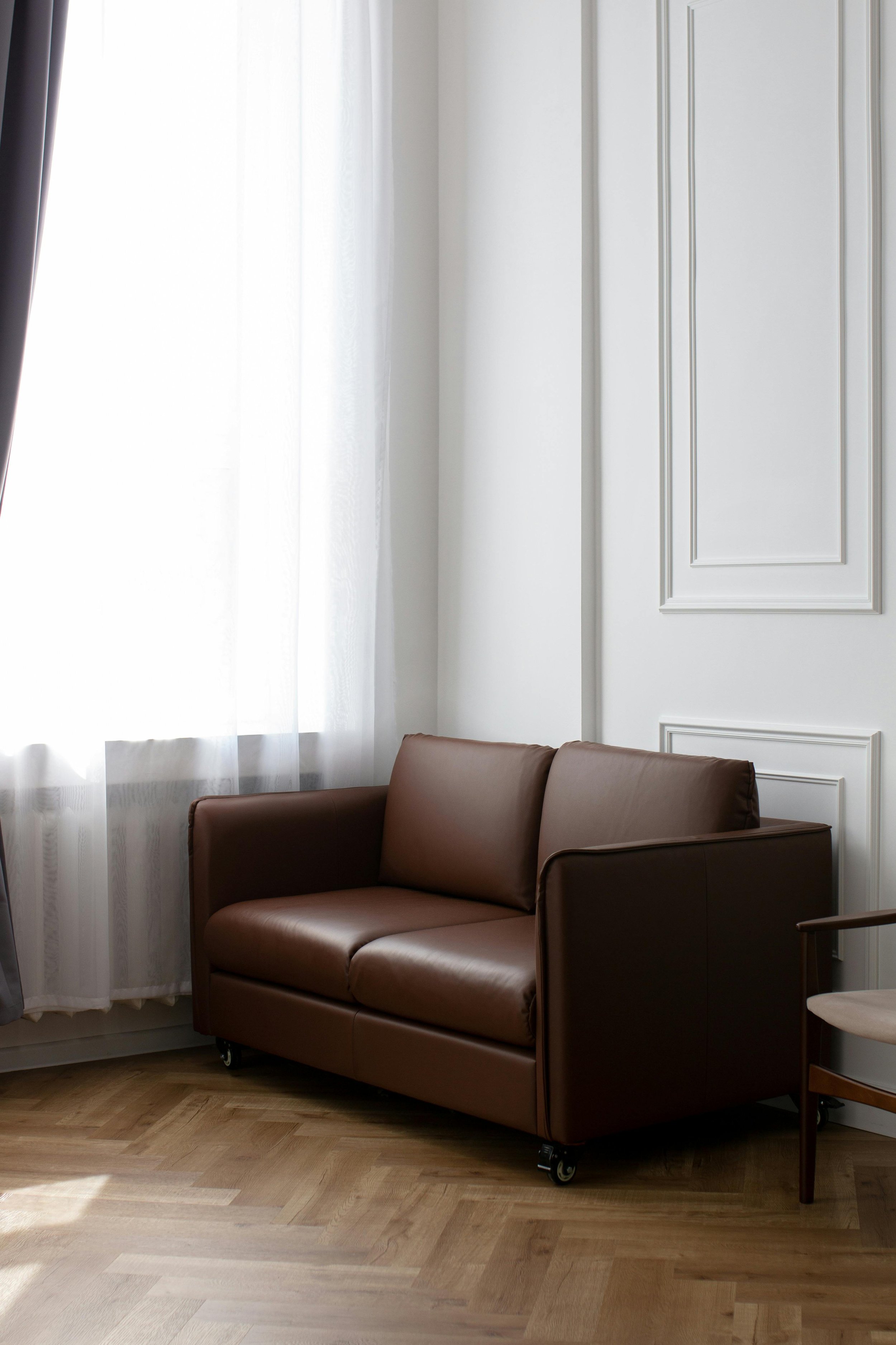 A small brown leather sofa with wheels in a well-lit room near a window with sheer white curtains and a chair beside it.