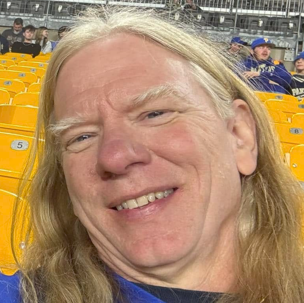 A smiling man with long blond hair at a sports stadium with yellow seats and other spectators in the background.