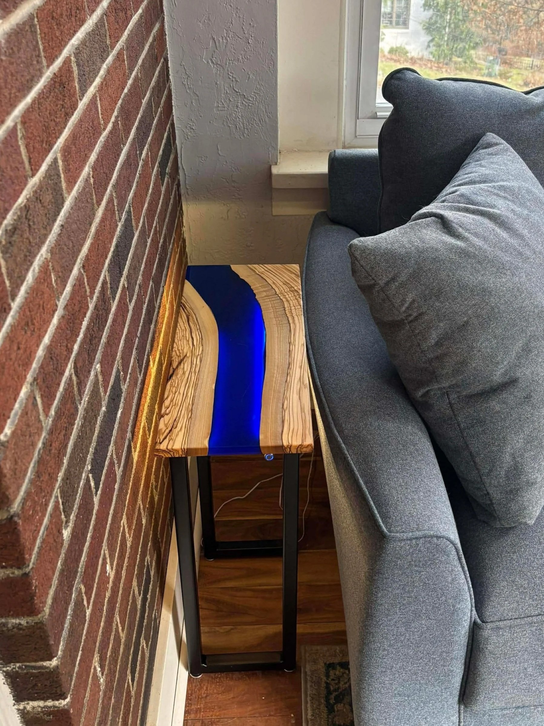 Wooden table with blue resin river design, next to a gray sofa with pillows, near a brick wall and windows.