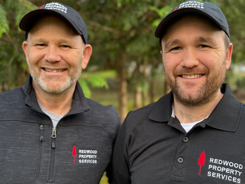 Two smiling men wearing black caps and jackets with 'Redwood Property Services' logo standing outdoors with trees in the background.