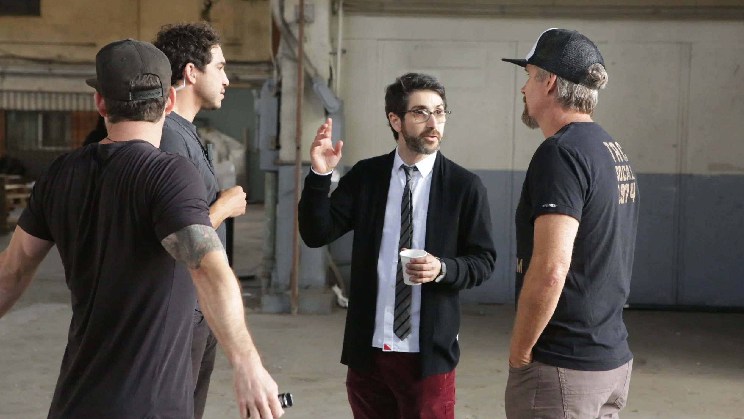 Four men engaged in a conversation inside an industrial-style space. One man with glasses, a beard, in a white shirt, black jacket, and striped tie is gesturing with his right hand and holding a coffee mug. The other three men are wearing casual black t-shirts and hats, listening to him.