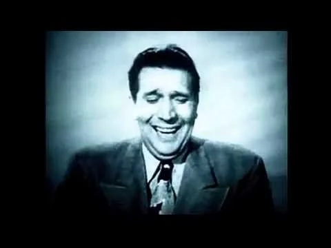 Black and white image of a man smiling and laughing, wearing a suit and tie.