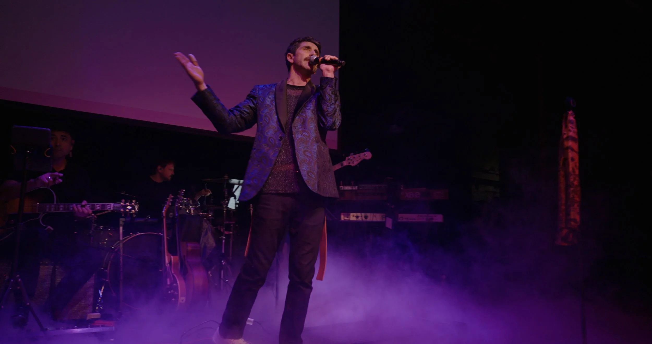 Male singer in a blue patterned blazer performing on stage with purple lighting, accompanied by musicians playing guitars and drums amid stage fog.
