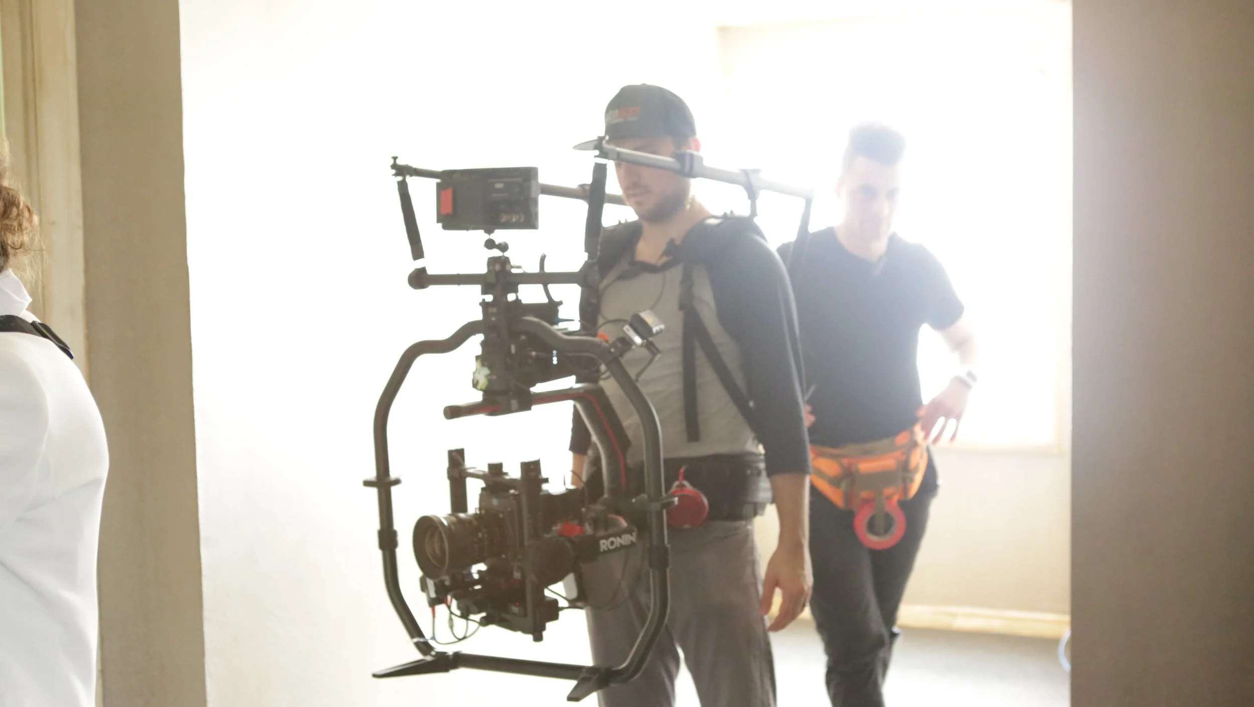 Filmmaker using a camera stabilizer rig during a shoot in a brightly lit room, with another crew member standing in the background.
