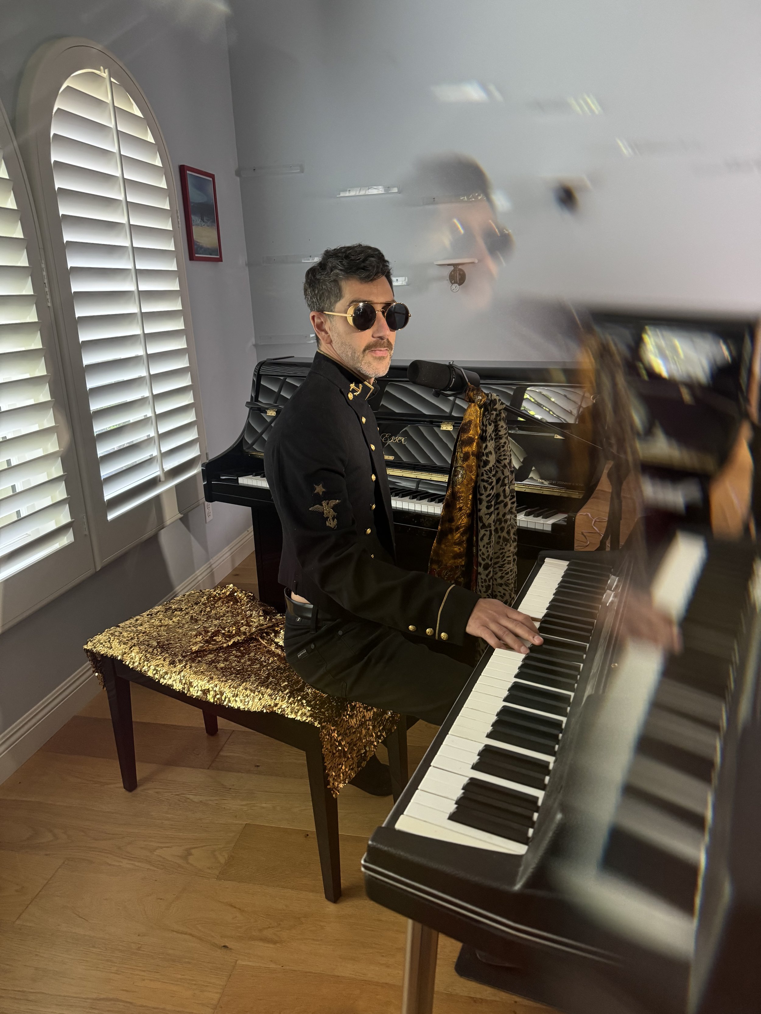 A man wearing sunglasses and a black military-style jacket is sitting at a piano, playing it. The room has white walls, a window with shutters, and a picture on the wall. There is a sequined gold-colored cloth on the piano bench.