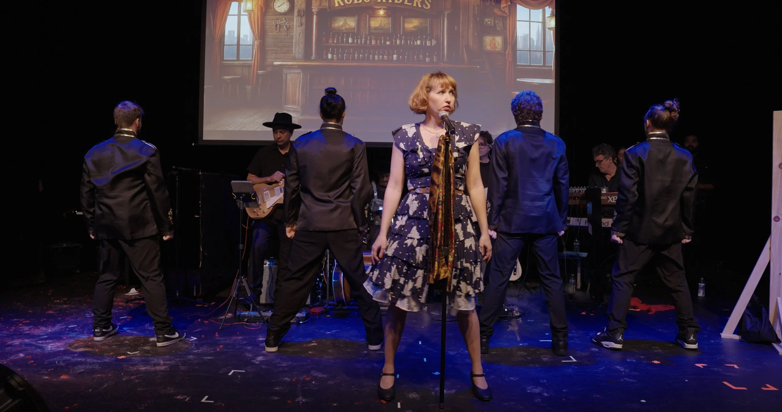 A stage performance featuring a woman in a patterned dress and black heels standing in front of a microphone, with her back to five dancers dressed in black suits and a band playing instruments behind her. A large screen above shows a vintage bar sce