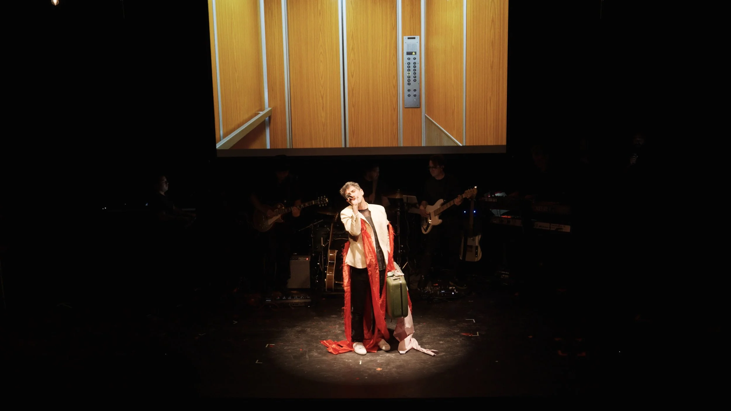 A man singing on stage with a suitcase, with a band playing guitars and keyboards behind him, and a projection of an elevator interior on the screen above him.