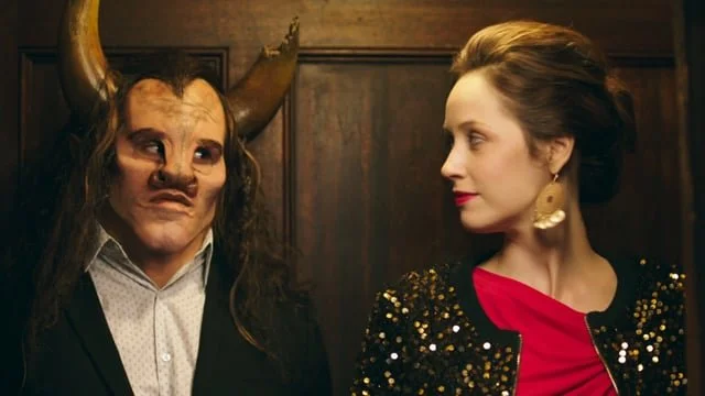 A person dressed as a devil with horns and makeup standing next to a woman with earrings and styled hair in an indoor setting.