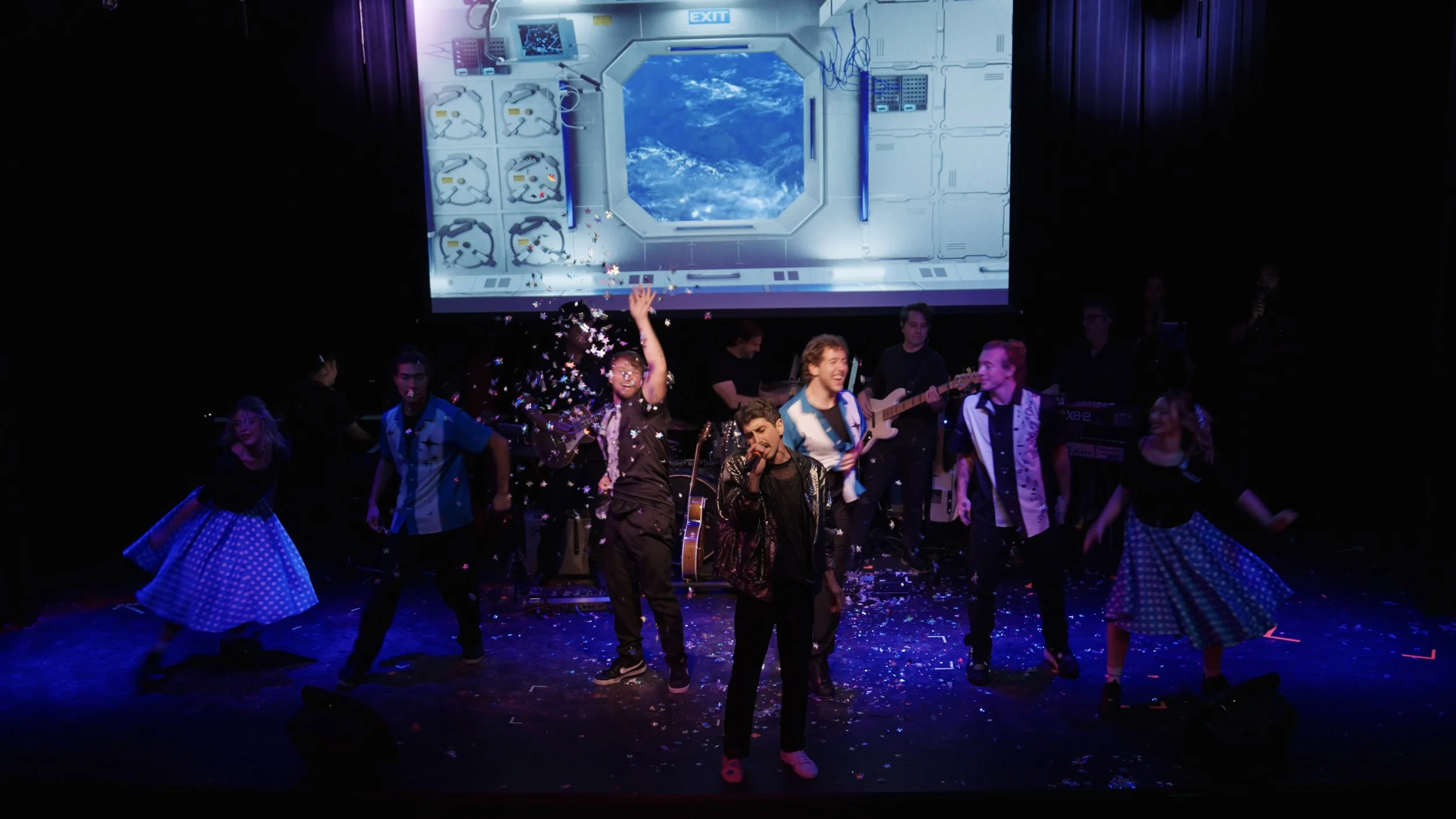 A group of performers on stage with musical instruments, singing and dancing, with a large screen showing space station imagery in the background and confetti falling around.