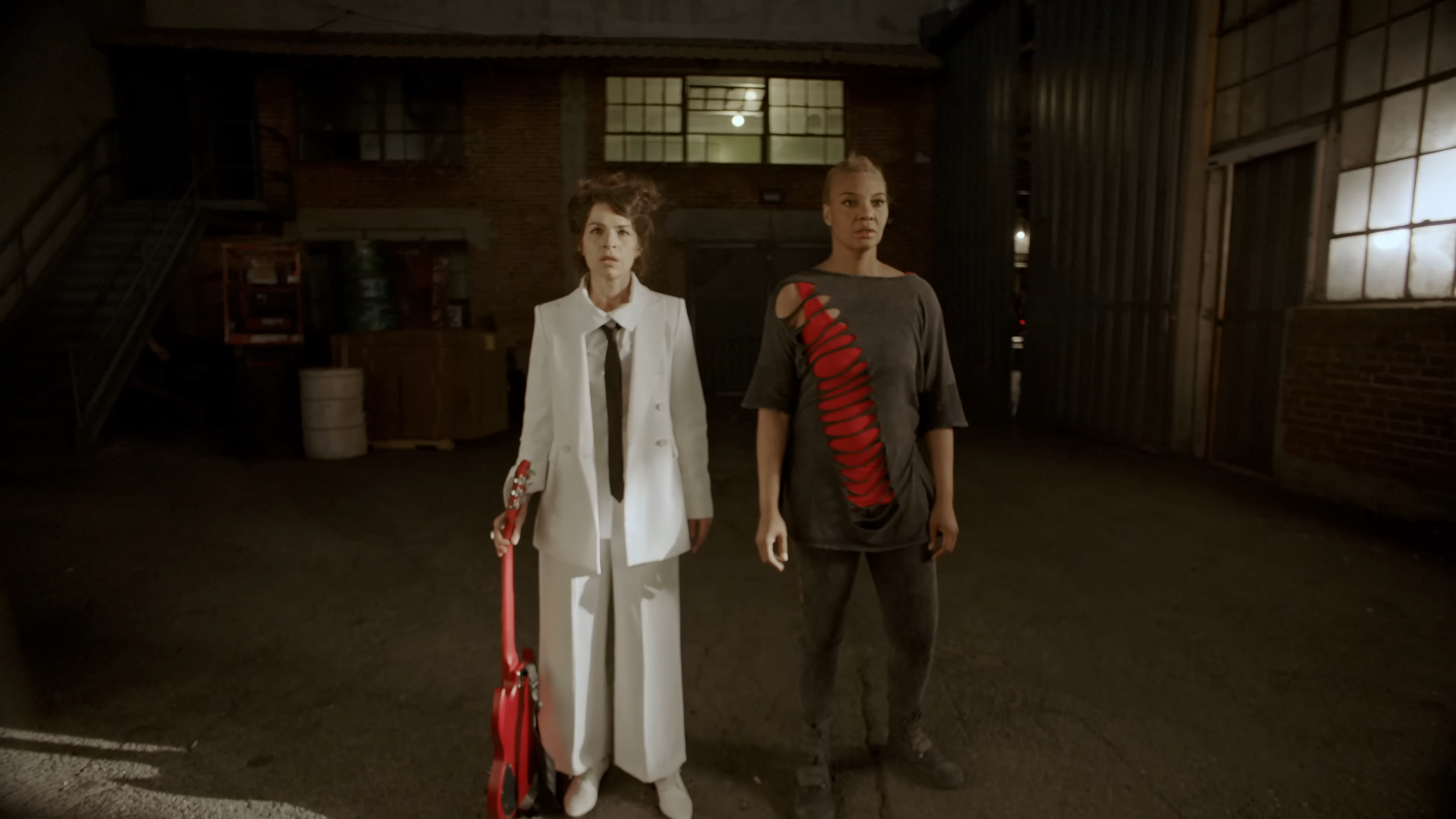 Two women standing in a dimly lit industrial warehouse. One is dressed in a white suit with black tie holding a red leash, and the other is wearing a distressed gray T-shirt with red slash design and black pants.