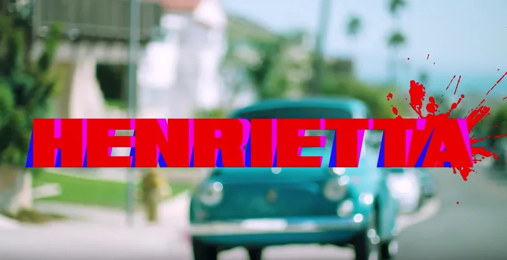 Blurred photo of a vintage teal Volkswagen Beetle car on a street with a background of trees and houses. Overlaid text in bold red and blue says 'HENRIETTA' with a red splatter effect.