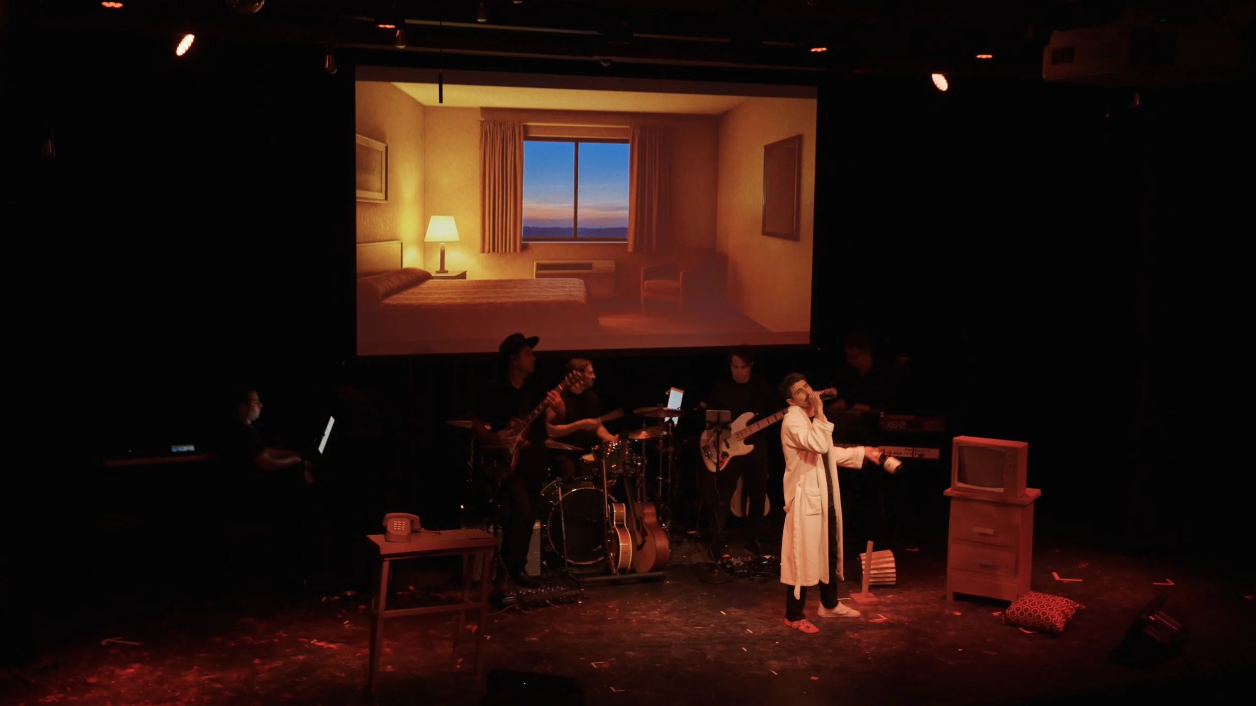 A band performs on stage with a large backdrop displaying a bedroom scene with a sunset view outside the window.