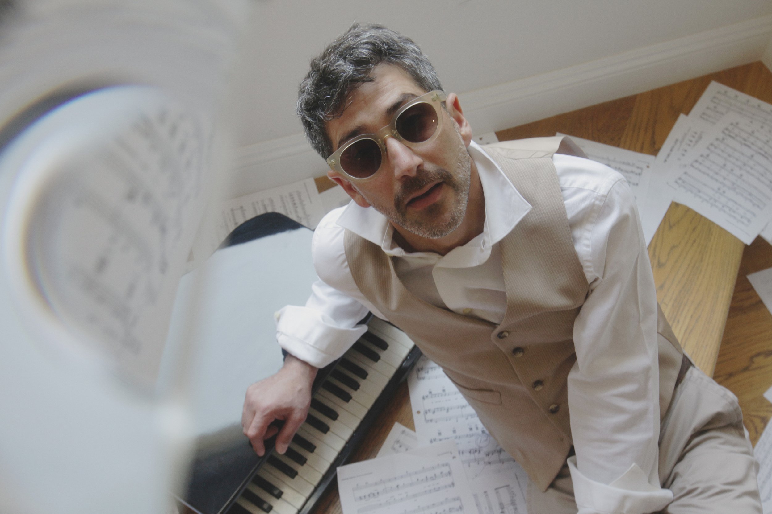 Man with sunglasses and gray hair lying on floor with scattered sheet music and a small keyboard, looking up at camera.