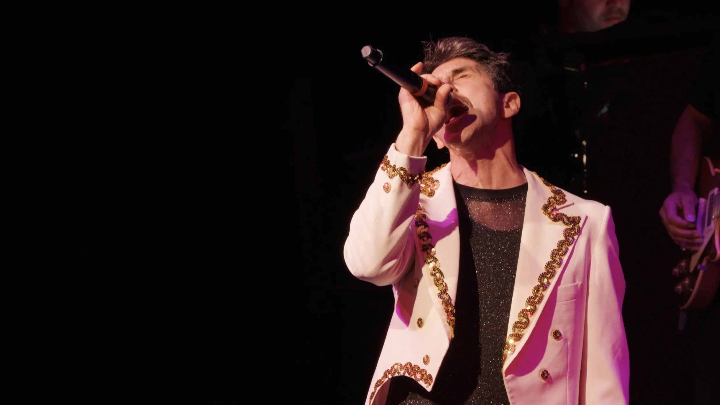 A male singer passionately singing into a microphone, wearing an ornate white jacket with gold embellishments and a sparkly black shirt, on stage with a dark background.