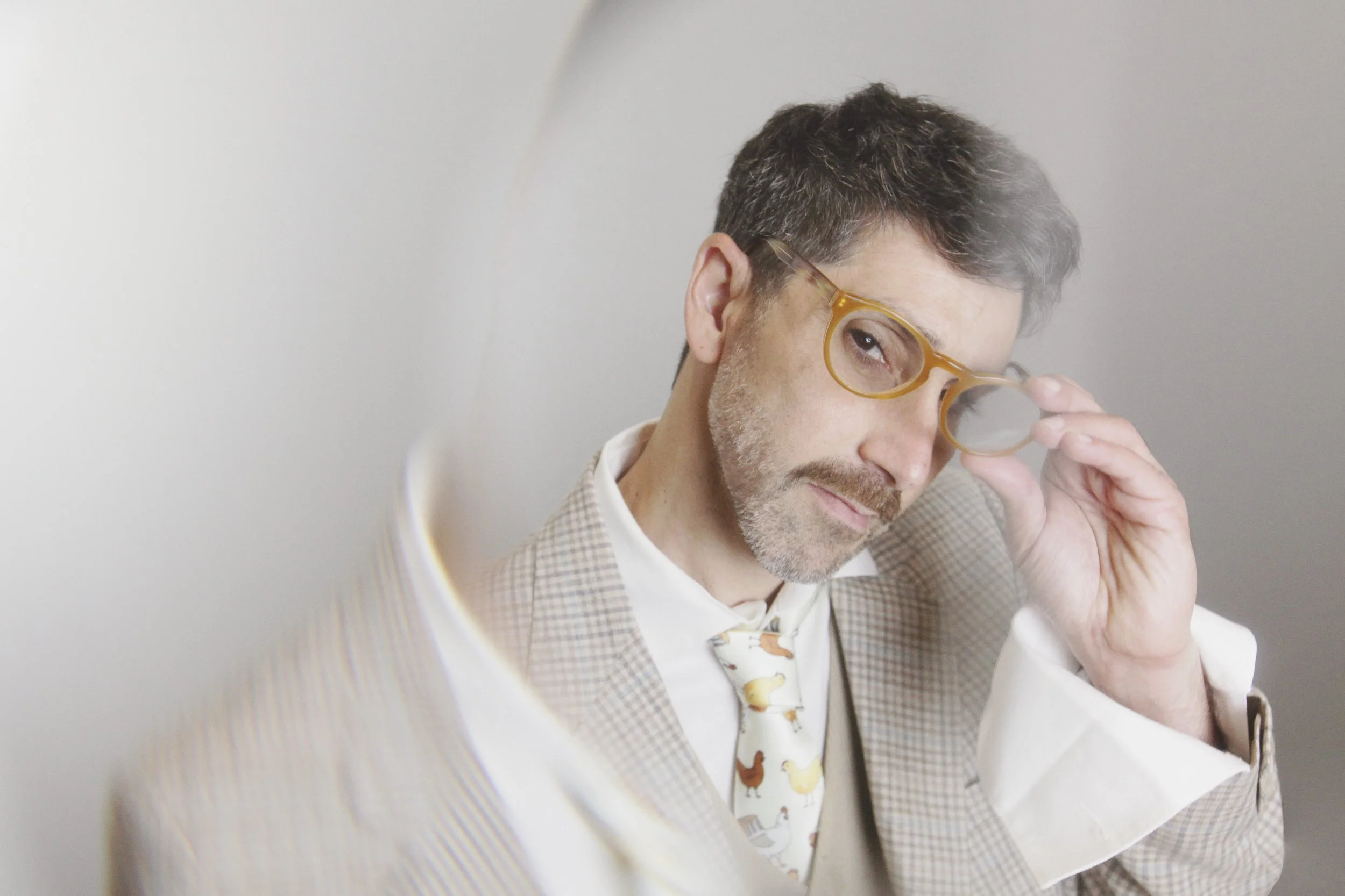 A man with dark hair, glasses, and a beard, adjusting his glasses with one hand, wearing a beige checkered blazer, white shirt, and a patterned tie with chickens, looking at the camera.