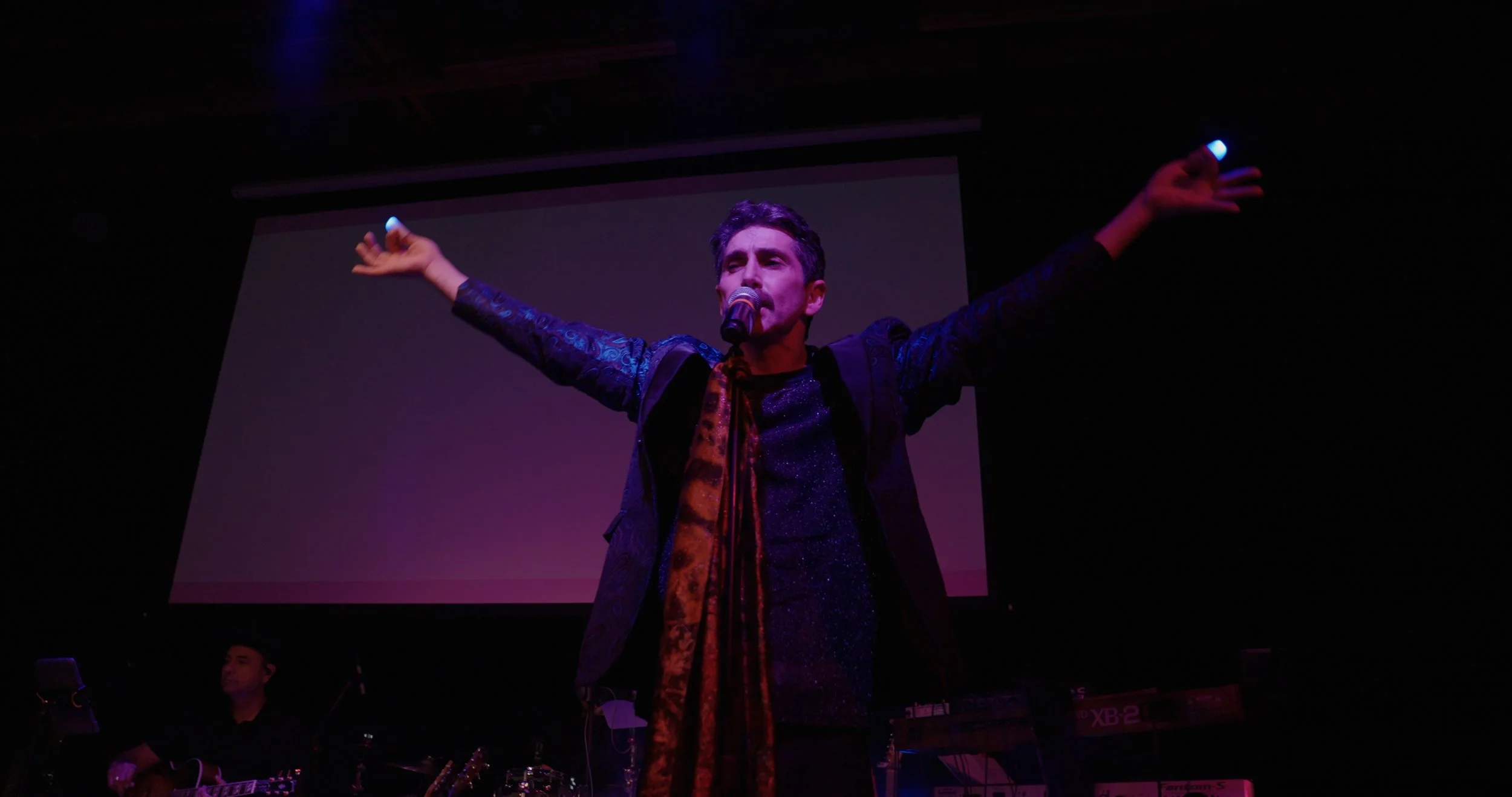 Performer singing on stage with arms outstretched and a microphone, with musicians and musical equipment in the background.