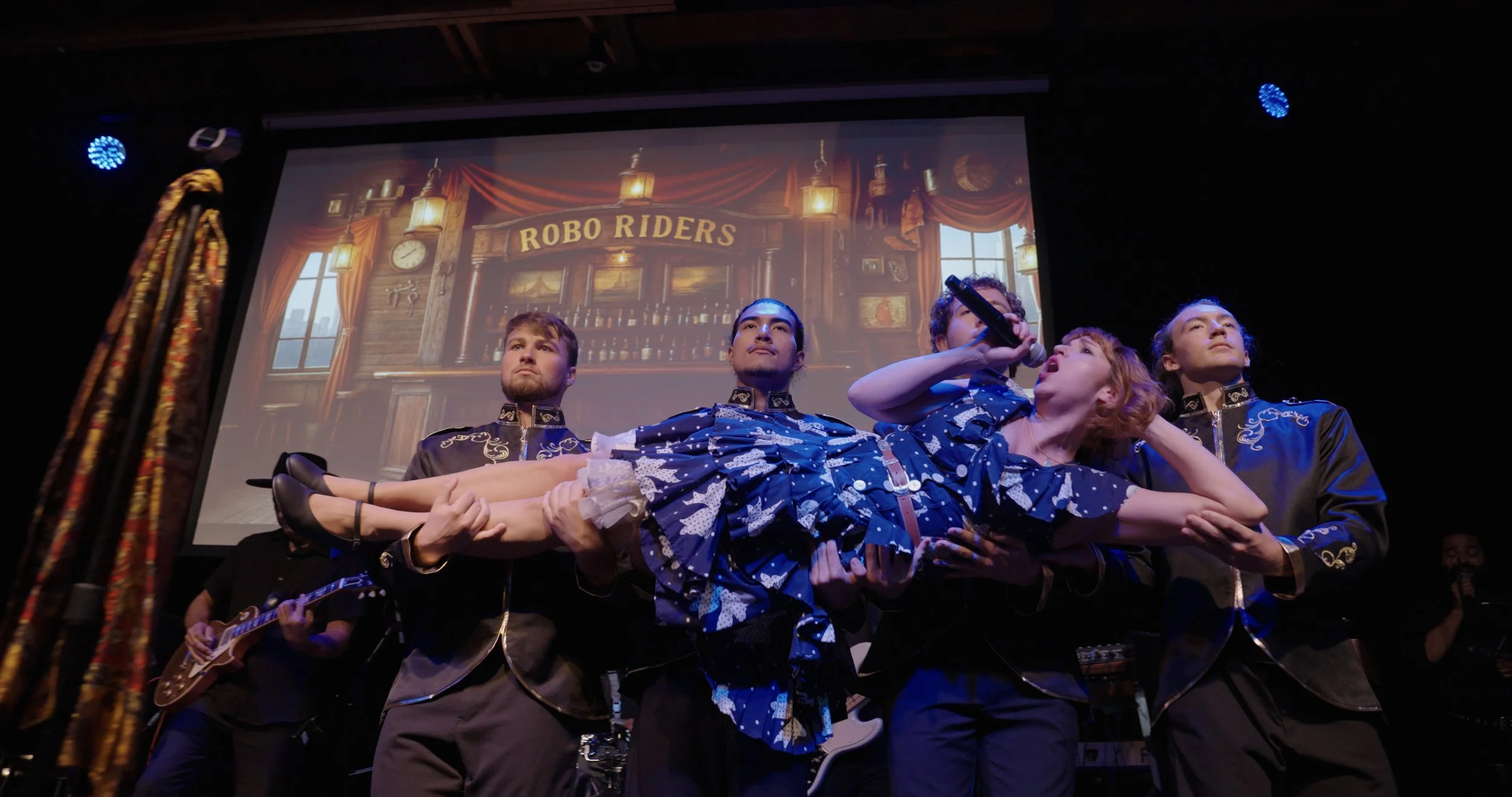 Stage performance with four performers lifting a woman in a blue dress, a scenic background showing a bar with a sign that reads 'Robo Riders', and a guitarist on the left, during a theatrical or musical show.
