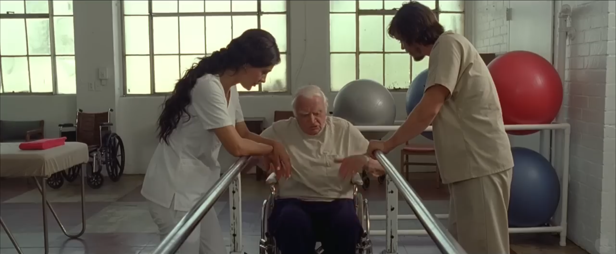 Two healthcare workers assist an elderly man in a wheelchair, helping him walk down a corridor in a medical or rehabilitation facility.