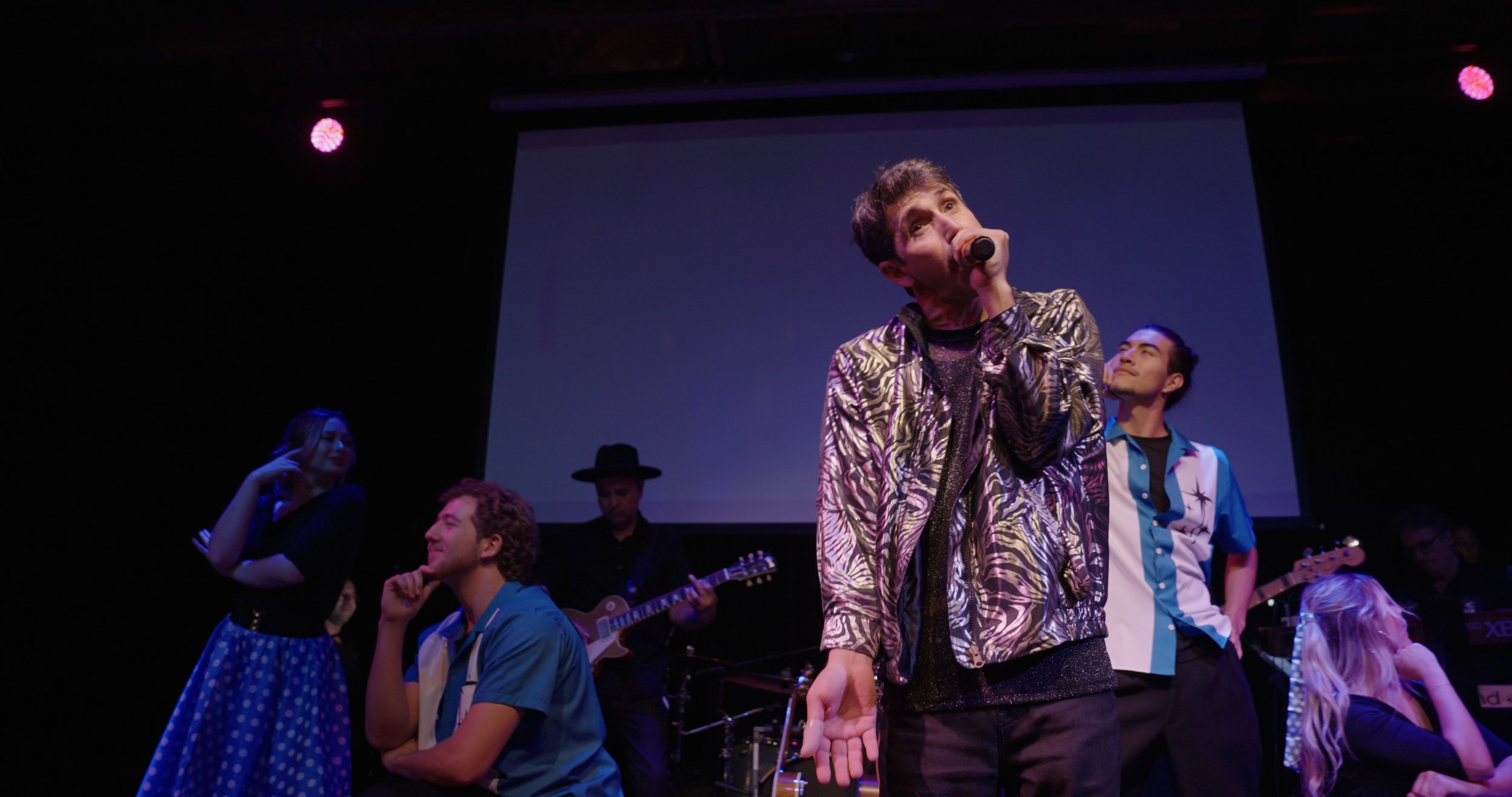 Group of performers on stage, with a man in the center holding a microphone, dressed in a patterned jacket, and others playing instruments and engaging. Scene is lit with stage lights, and a projection screen is visible in the background.