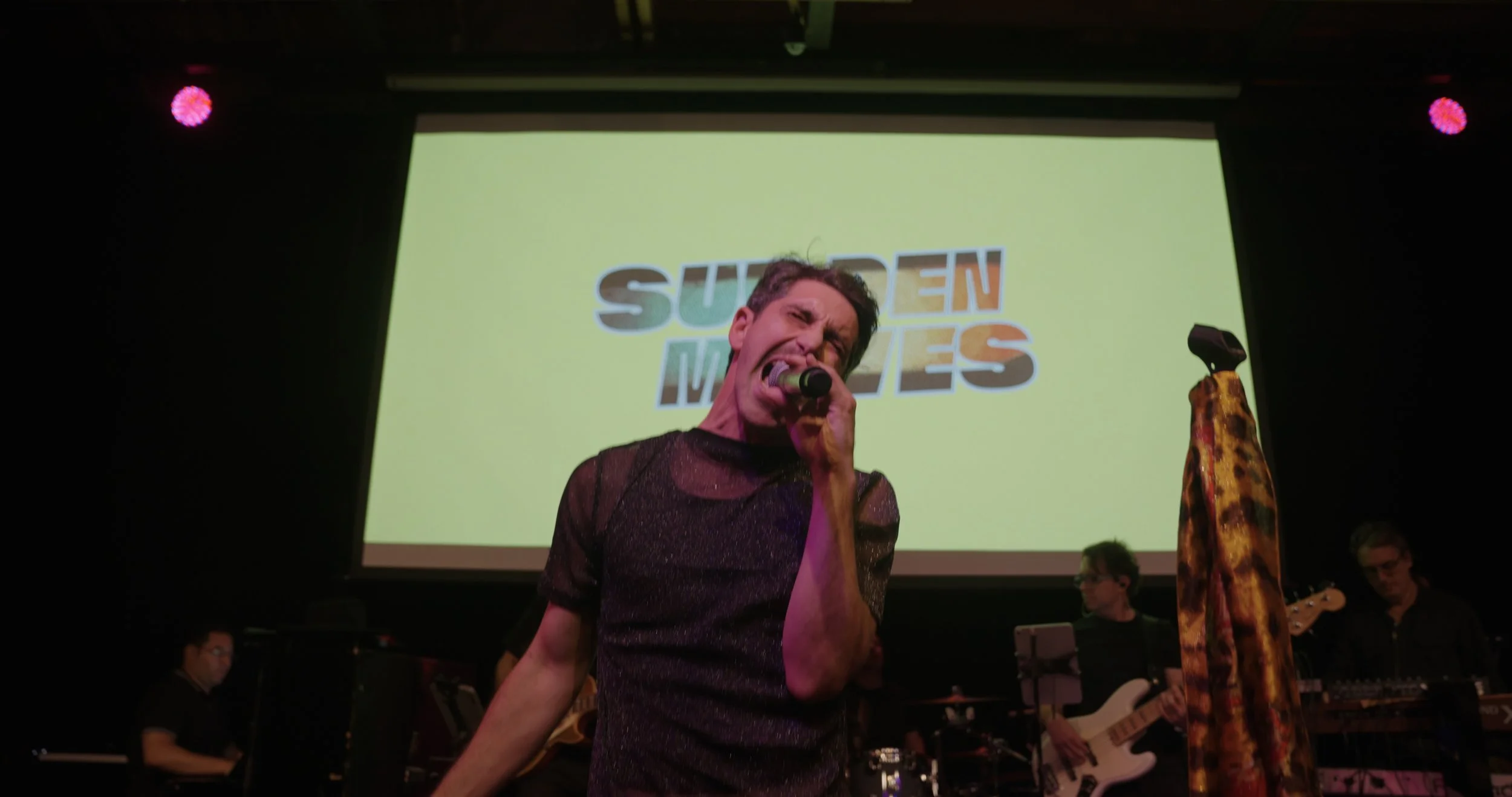 Man singing passionately on stage with a microphone, band members playing instruments in the background, large screen with 'SUDDEN MOVES' logo behind him, stage lighting includes pink spotlights.