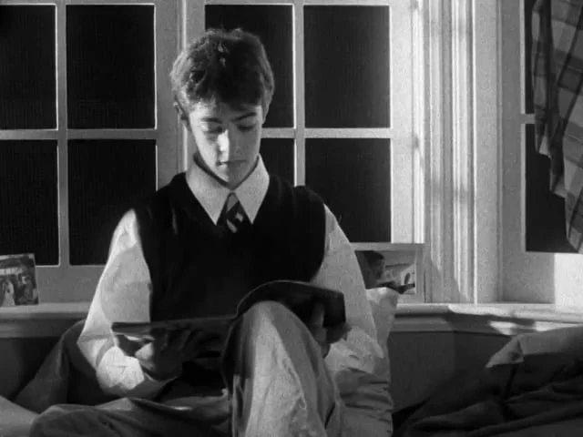 A young person sitting on a bed and reading a book in black and white.