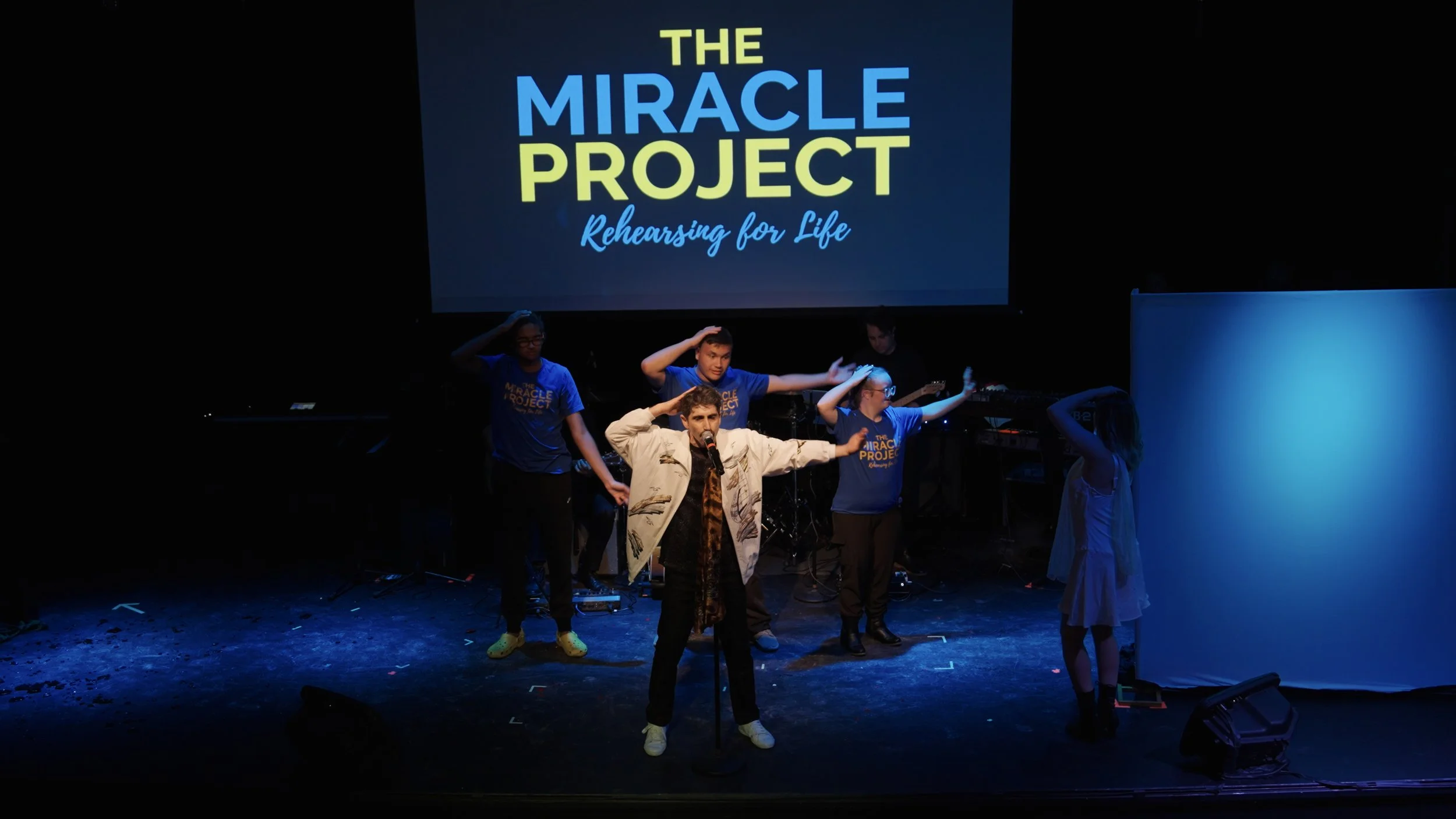 A stage performance for The Miracle Project with a singer in front and five people behind, some wearing matching blue T-shirts with the project logo, each saluting or with hands near their heads. A large screen above them displays the event's name an