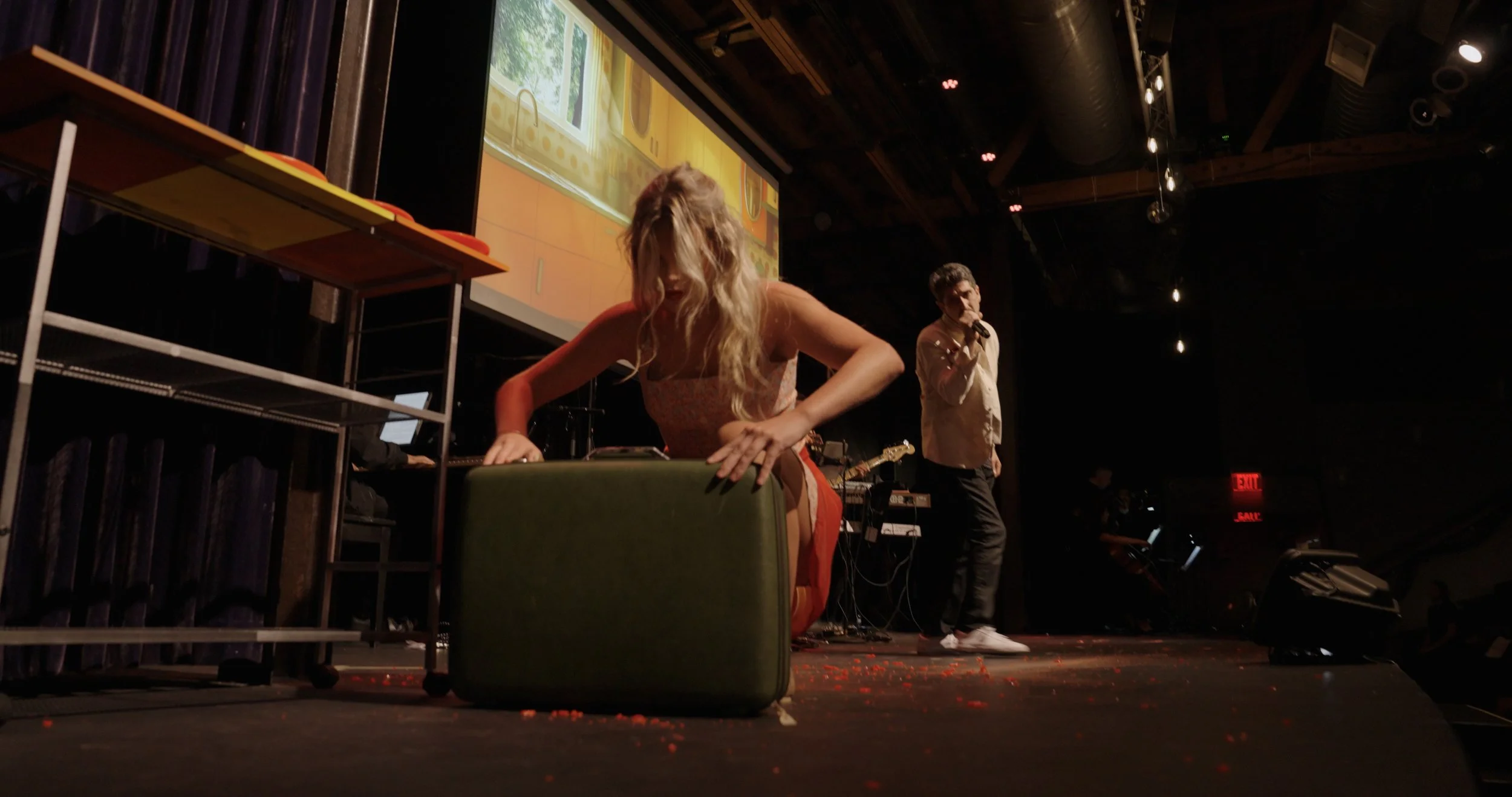 A woman with wavy blonde hair bending over on a stage, with her hand on a suitcase or box. A man standing with a microphone in the background, and another person near some equipment. There is a large screen behind them displaying a colorful cartoon o