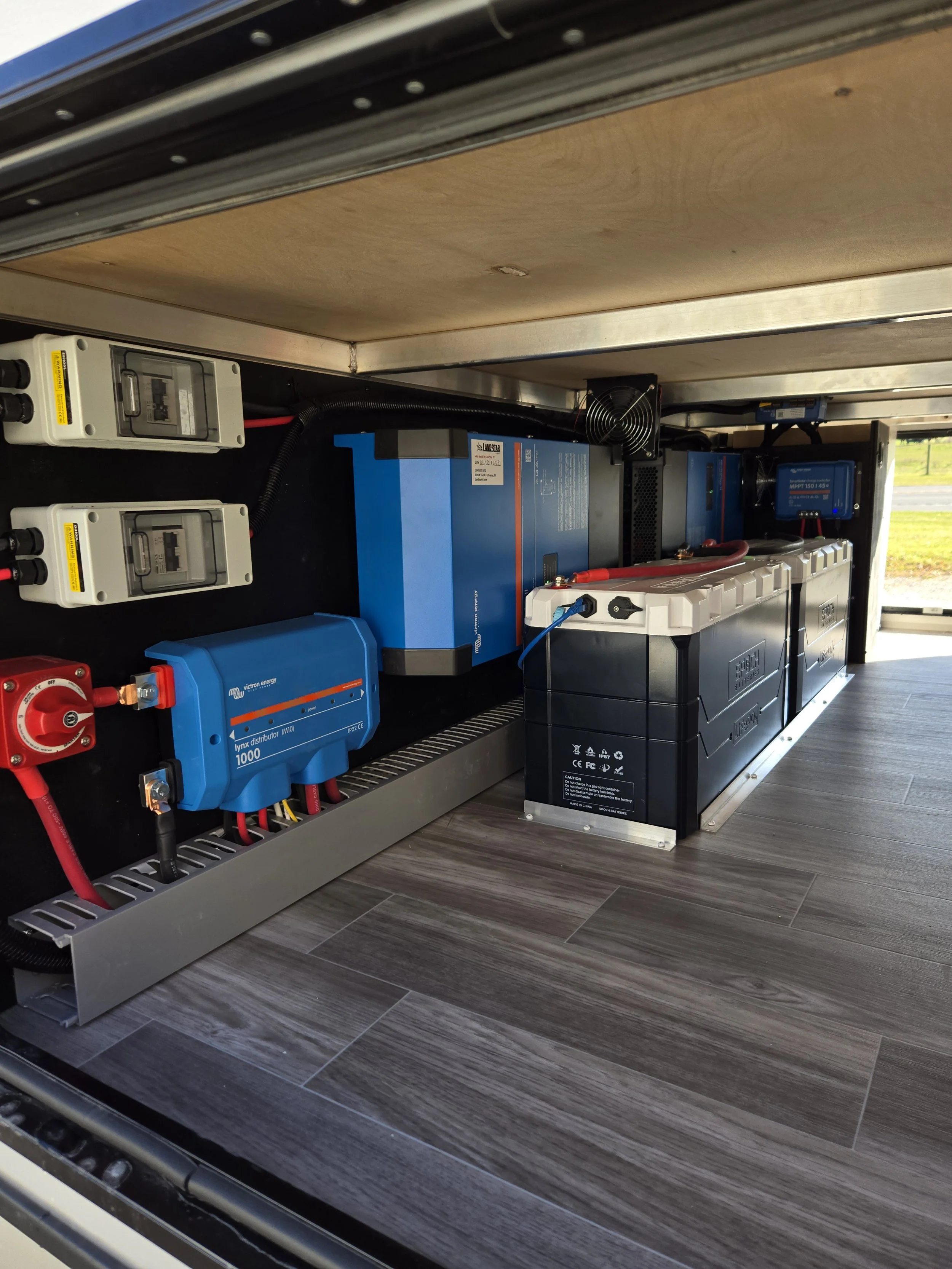 Inside of a technical cabinet showing batteries, inverters, and electrical components mounted on a black panel, with a wooden surface above and a gray floor below.