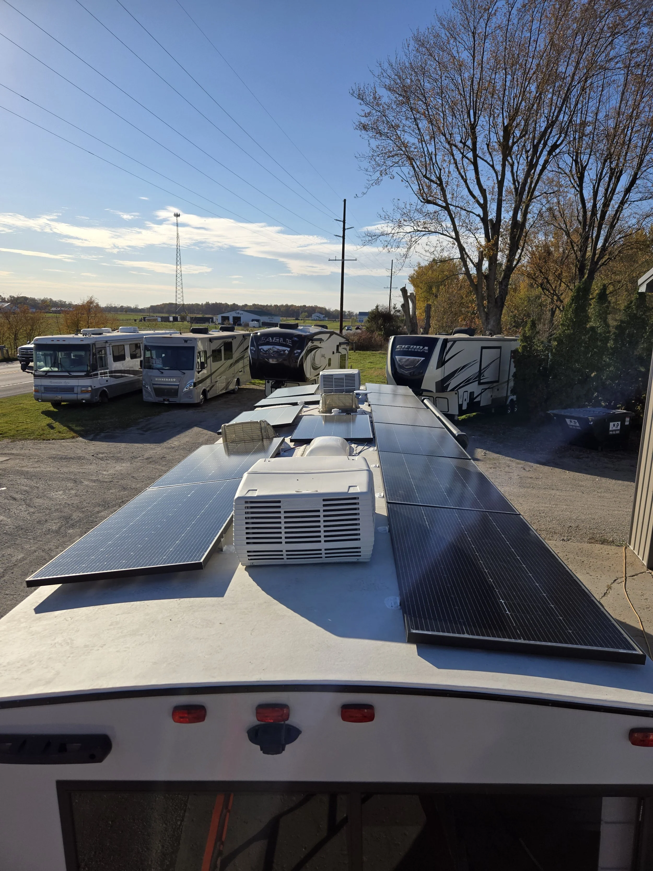 View from the roof of an RV with solar panels and an air conditioning unit, overlooking a parking lot with multiple RVs under a clear sky.