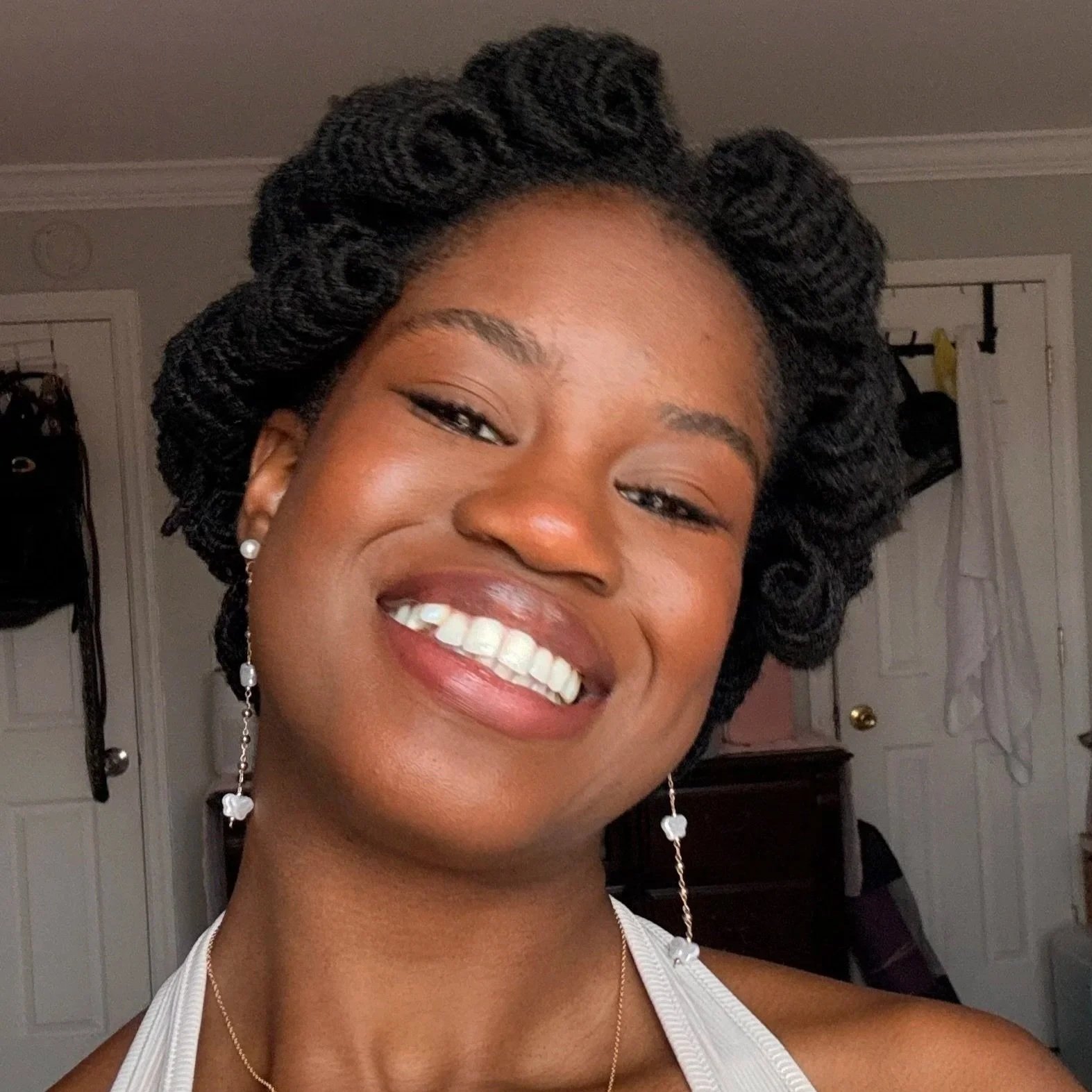 A young woman smiling with braided hair, wearing earrings and a sleeveless top, in a room with a closed door and hanging bags in the background.