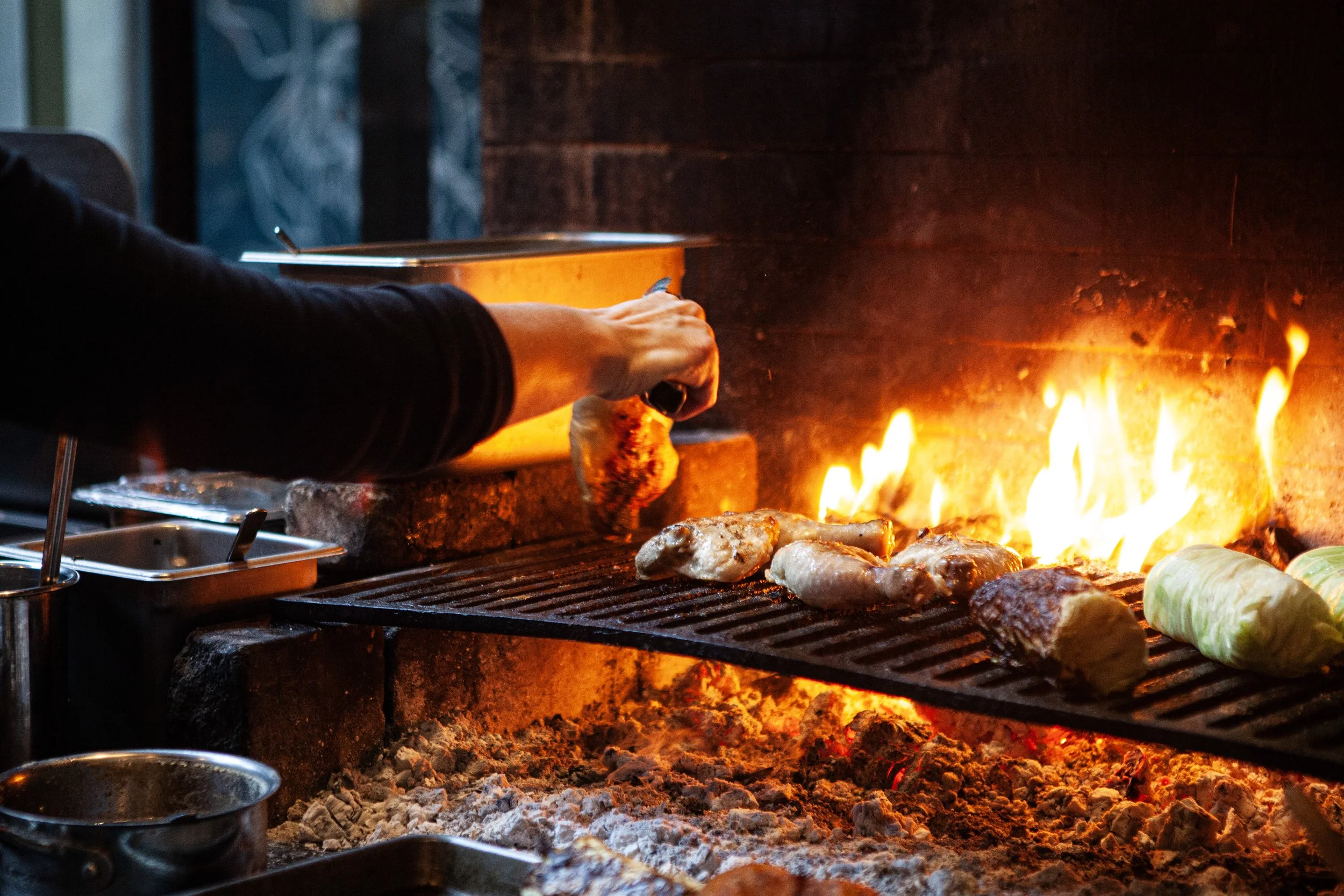 Person grilling chicken and vegetables over open flame in a wood-fired oven.
