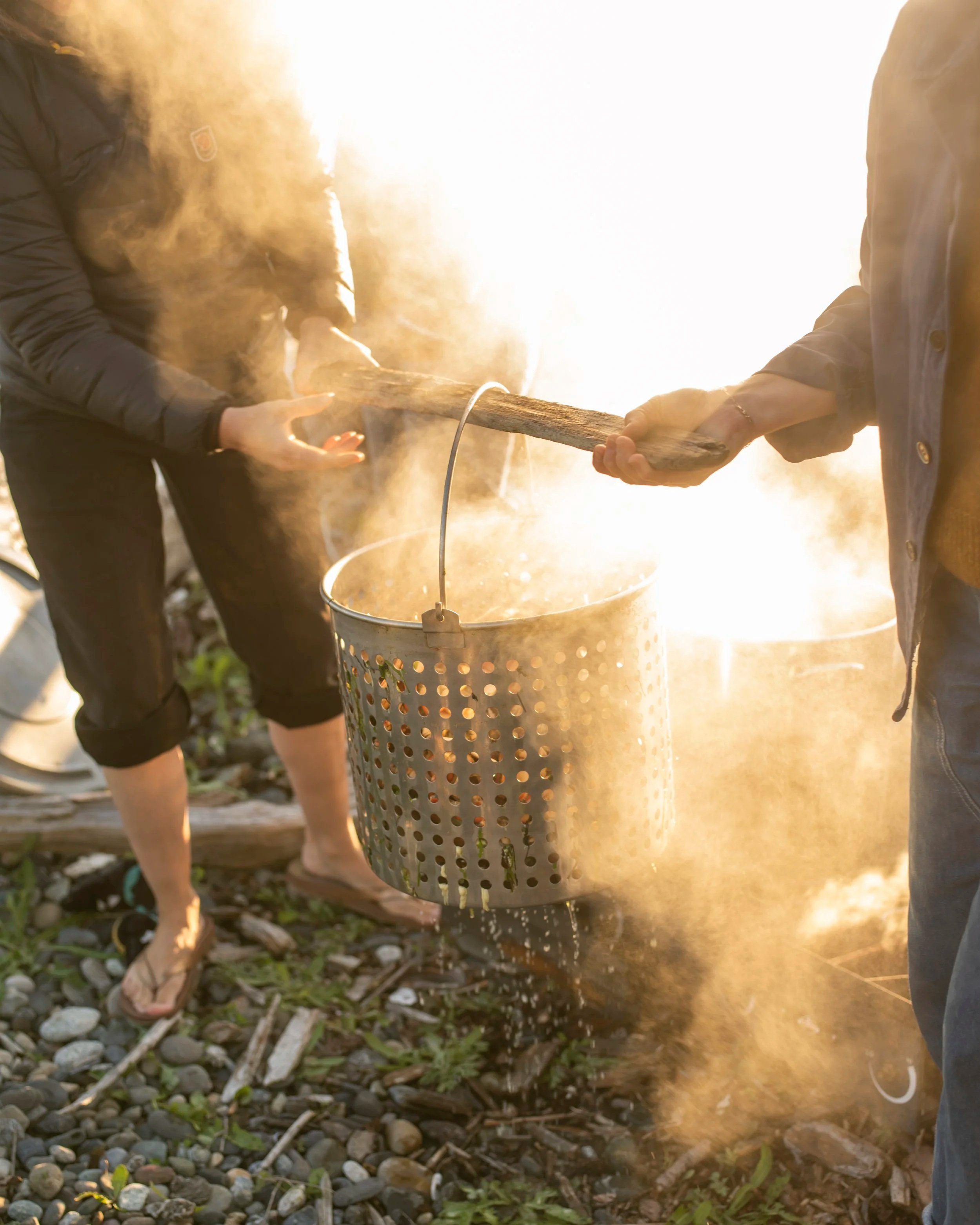 Two people holding a burning log over a metal bucket with smoke and sparks in the air, outdoors on a rocky ground.