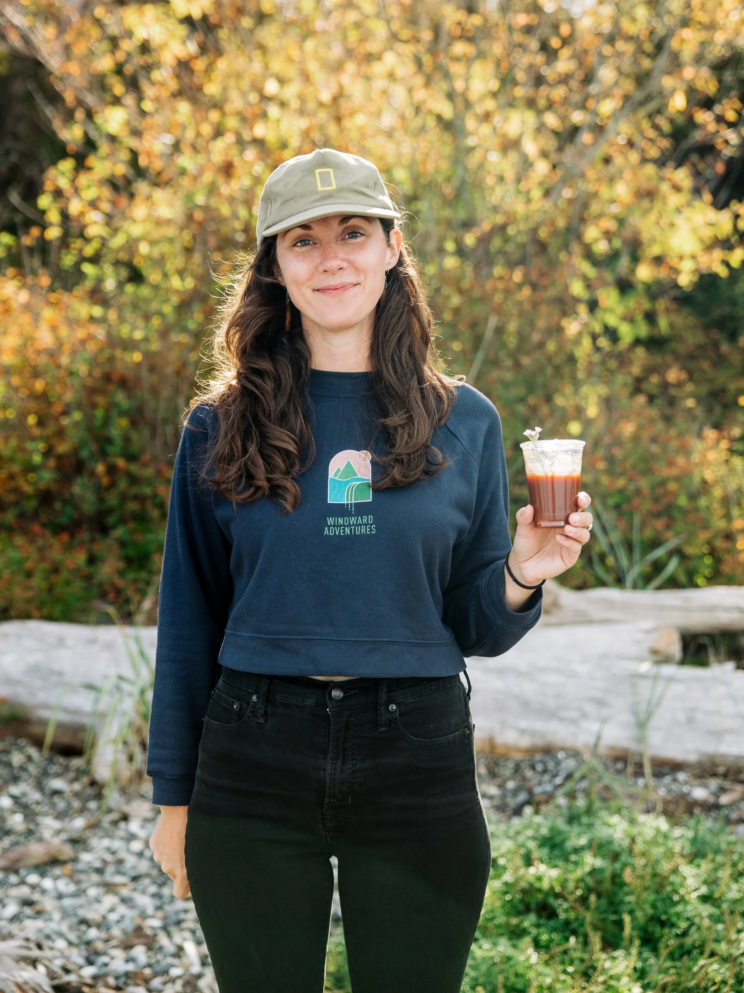 A woman with long wavy brown hair, wearing a beige cap and navy sweatshirt, holds a drink in a clear cup while standing outdoors in front of autumn foliage.