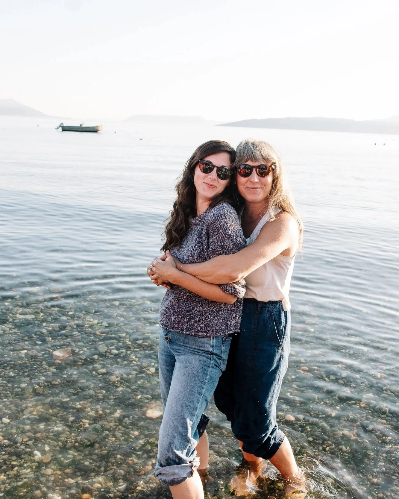 Two women standing in shallow water at the beach, embracing and smiling at the camera, with boats and distant islands in the background.