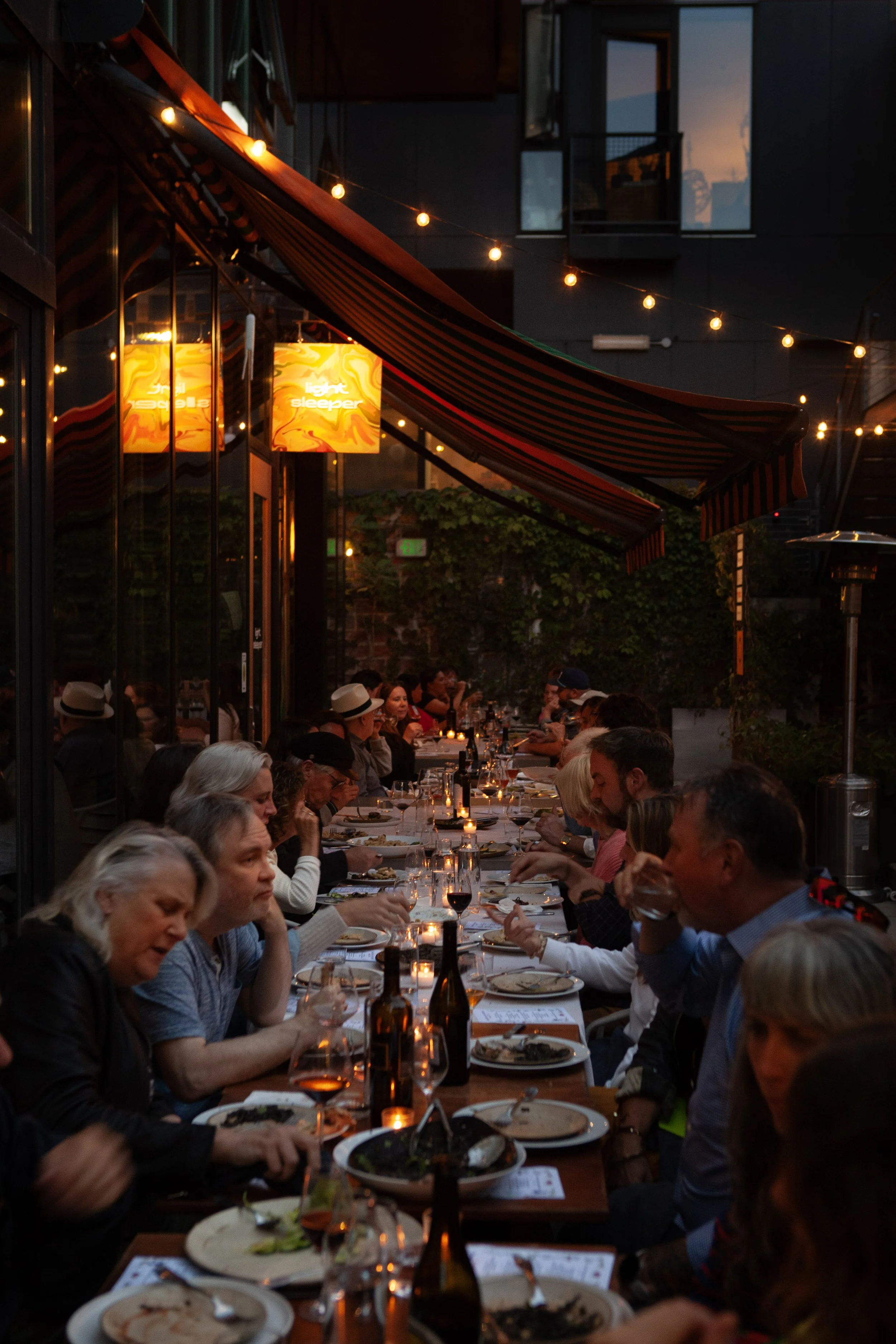 People dining outdoors at a long table during evening with string lights overhead and candles lit on the table.