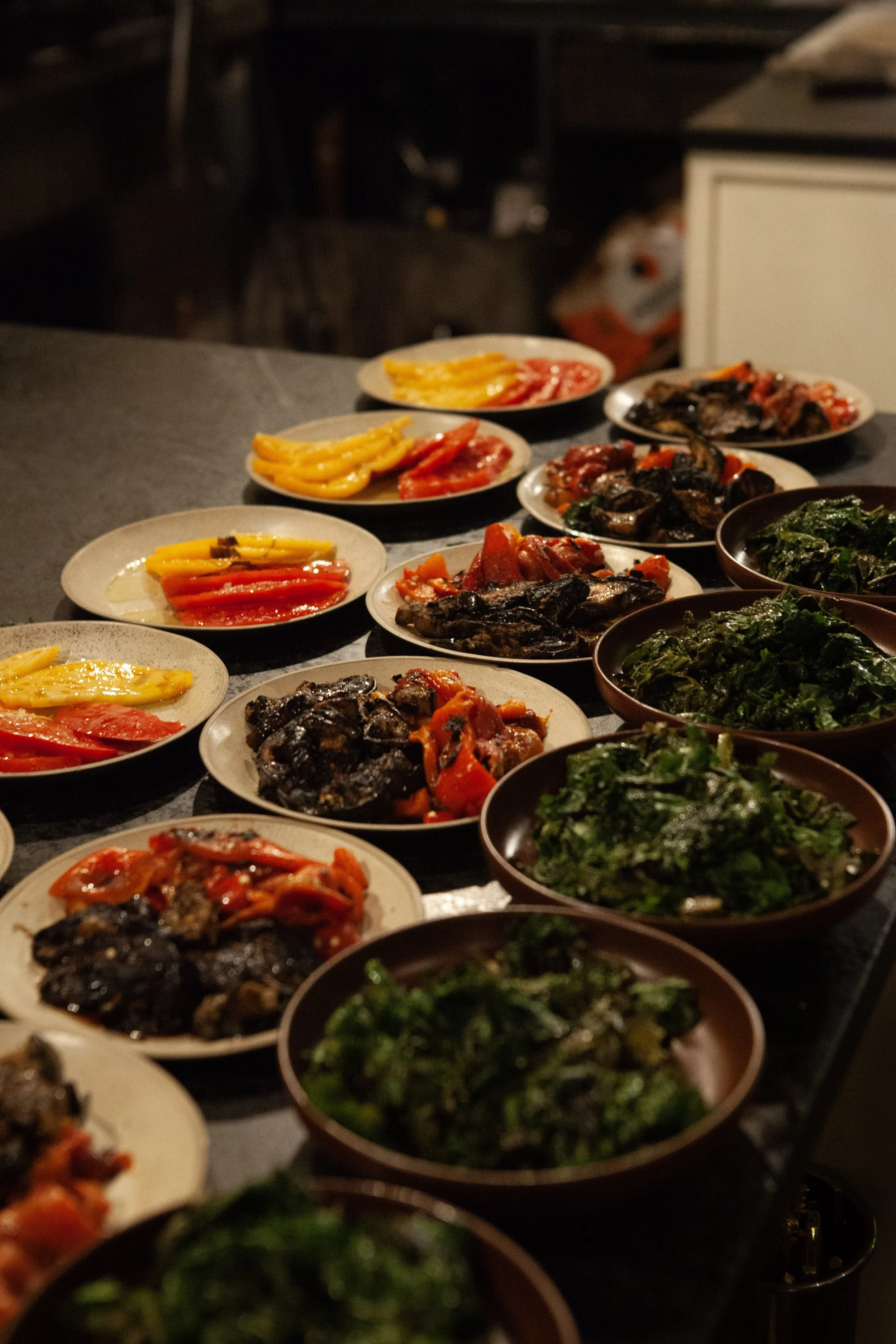 Various plates of roasted vegetables on a dark countertop, including peppers, eggplants, and greens.