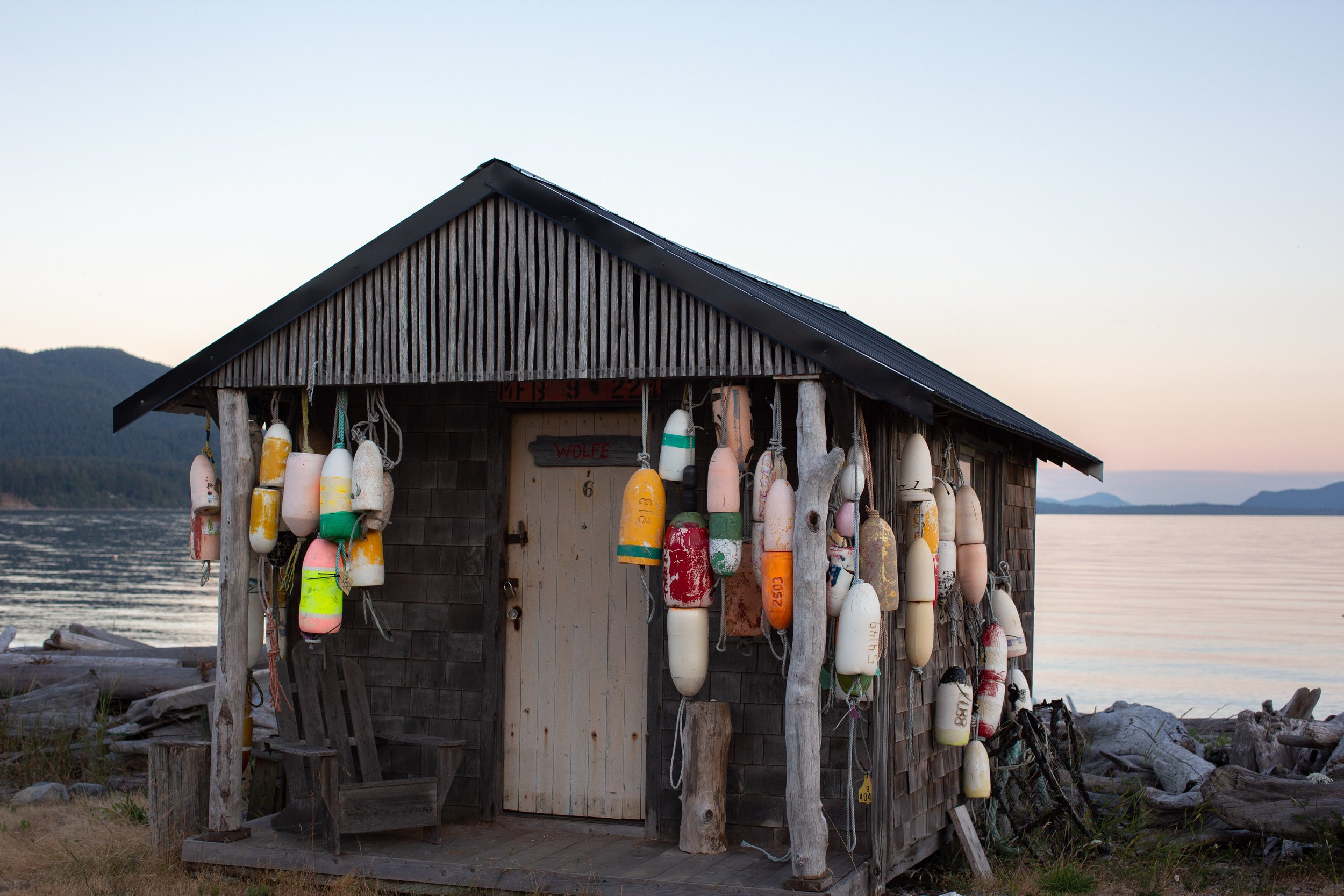 A small wooden boathouse by a lake, decorated with various colorful buoys hanging on the outside.