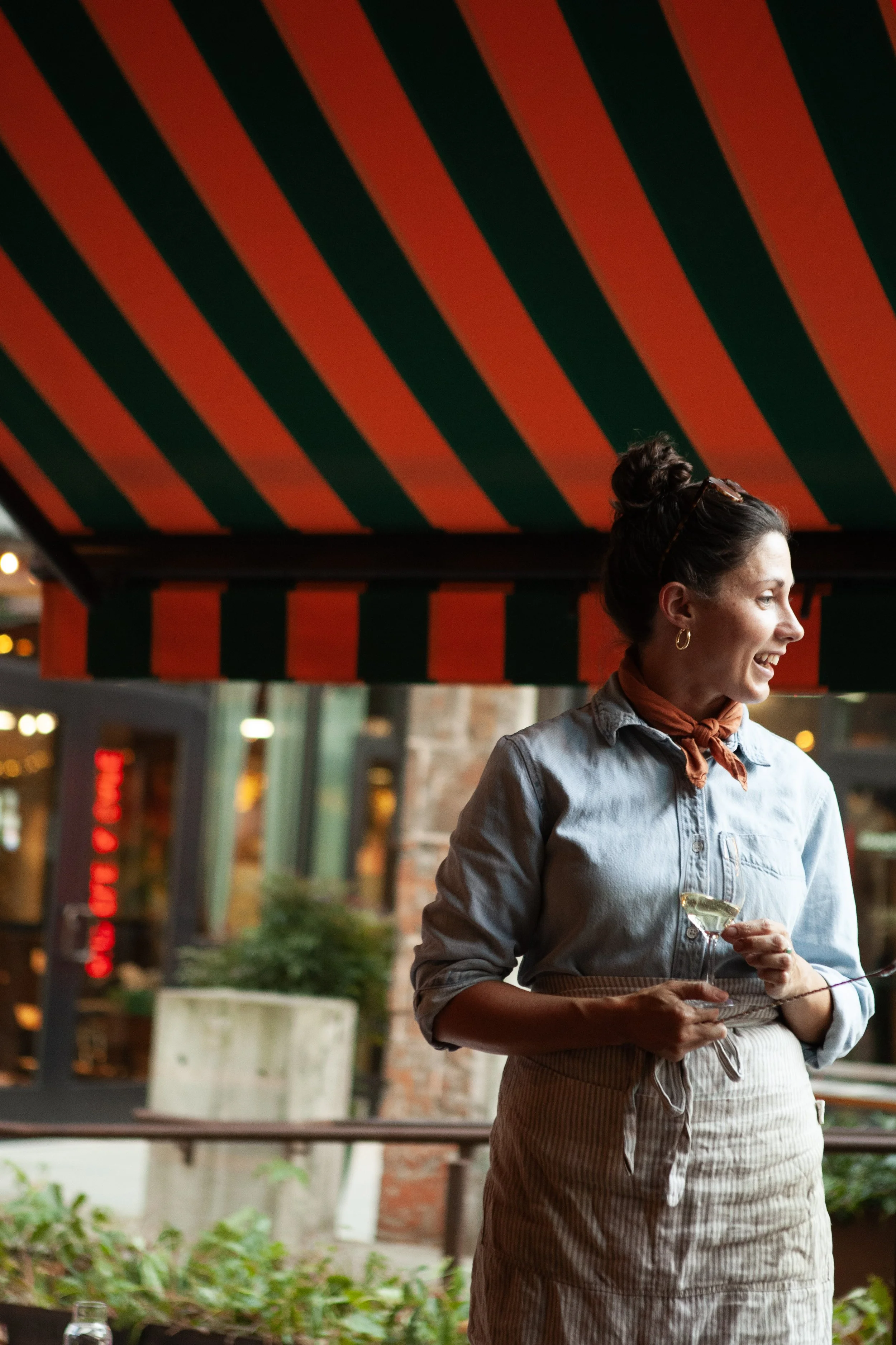 A woman with dark hair tied in a bun, wearing a light blue shirt, orange scarf, and striped apron, holding a cocktail glass, smiling and looking to her right under a red and green striped awning outdoors during evening.