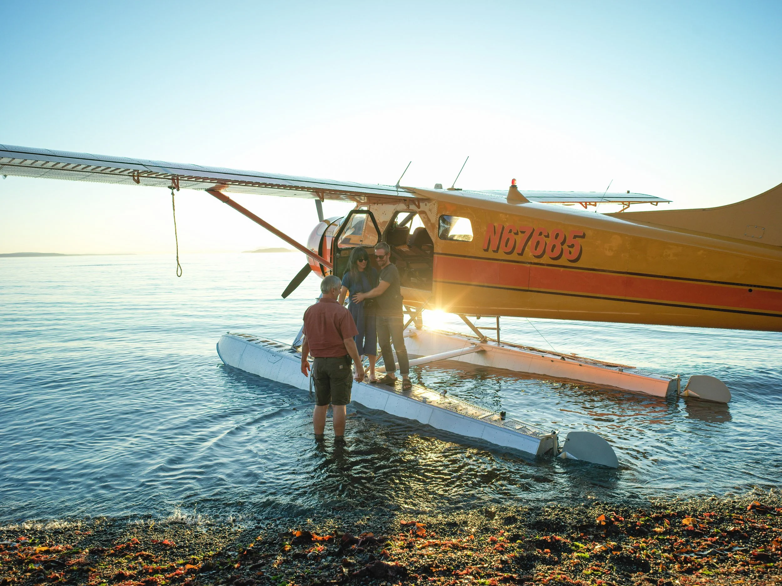 A seaplane on the water with two people boarding and another person standing in the water next to it, during sunset.