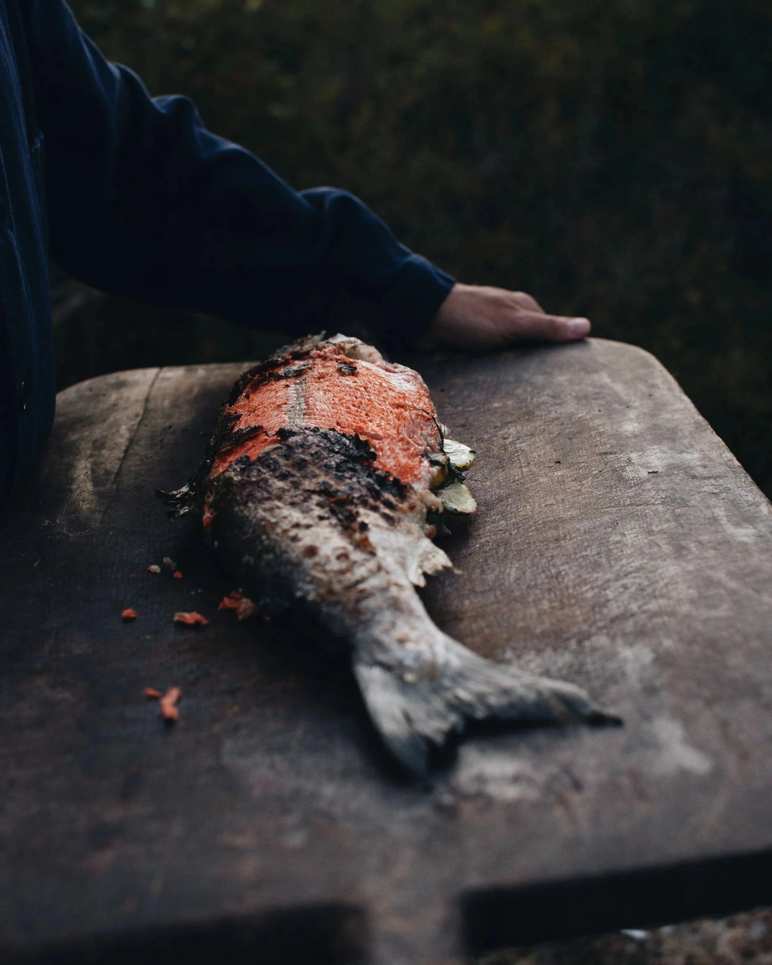 A cooked fish with orange and black scales on a wooden surface, with a person's hand resting nearby.