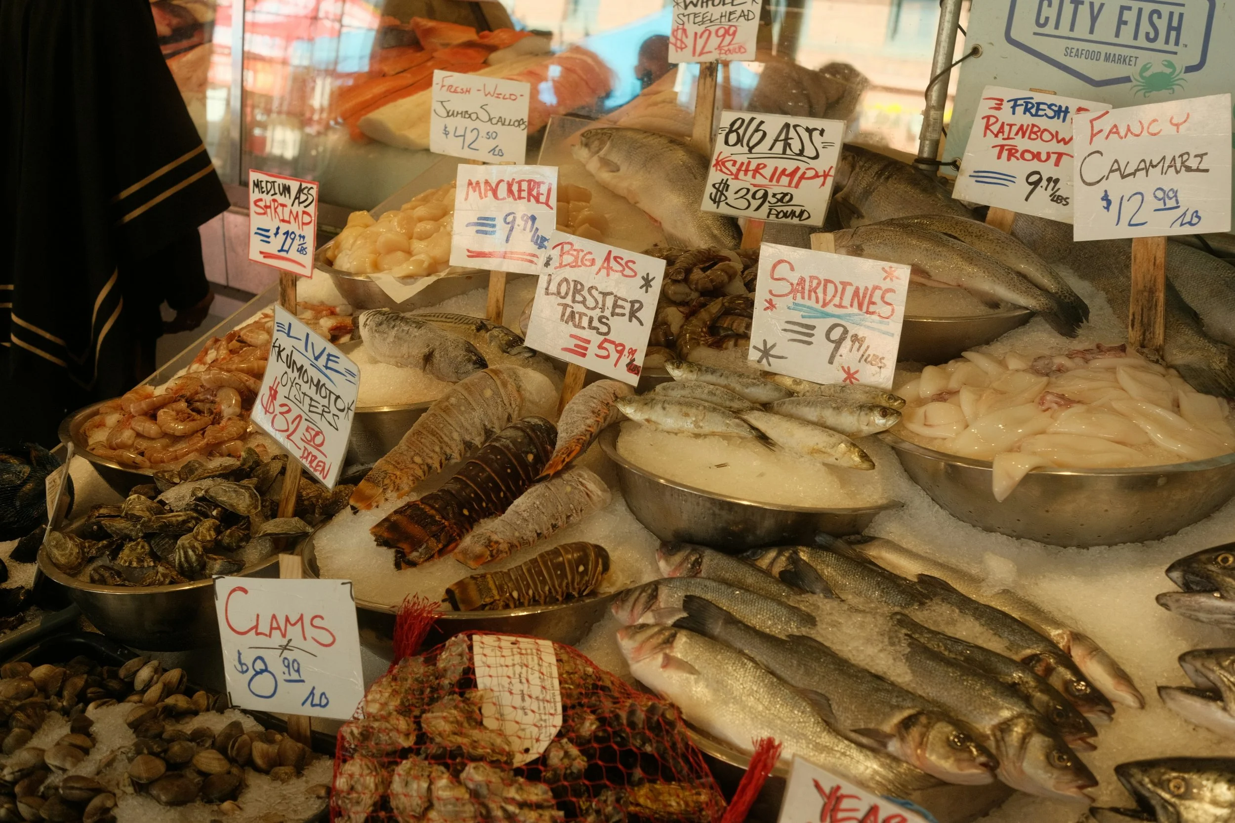 Fresh seafood display at a market, including fish, shrimp, oysters, squid, clams, and lobster tails, with handwritten price and product labels.