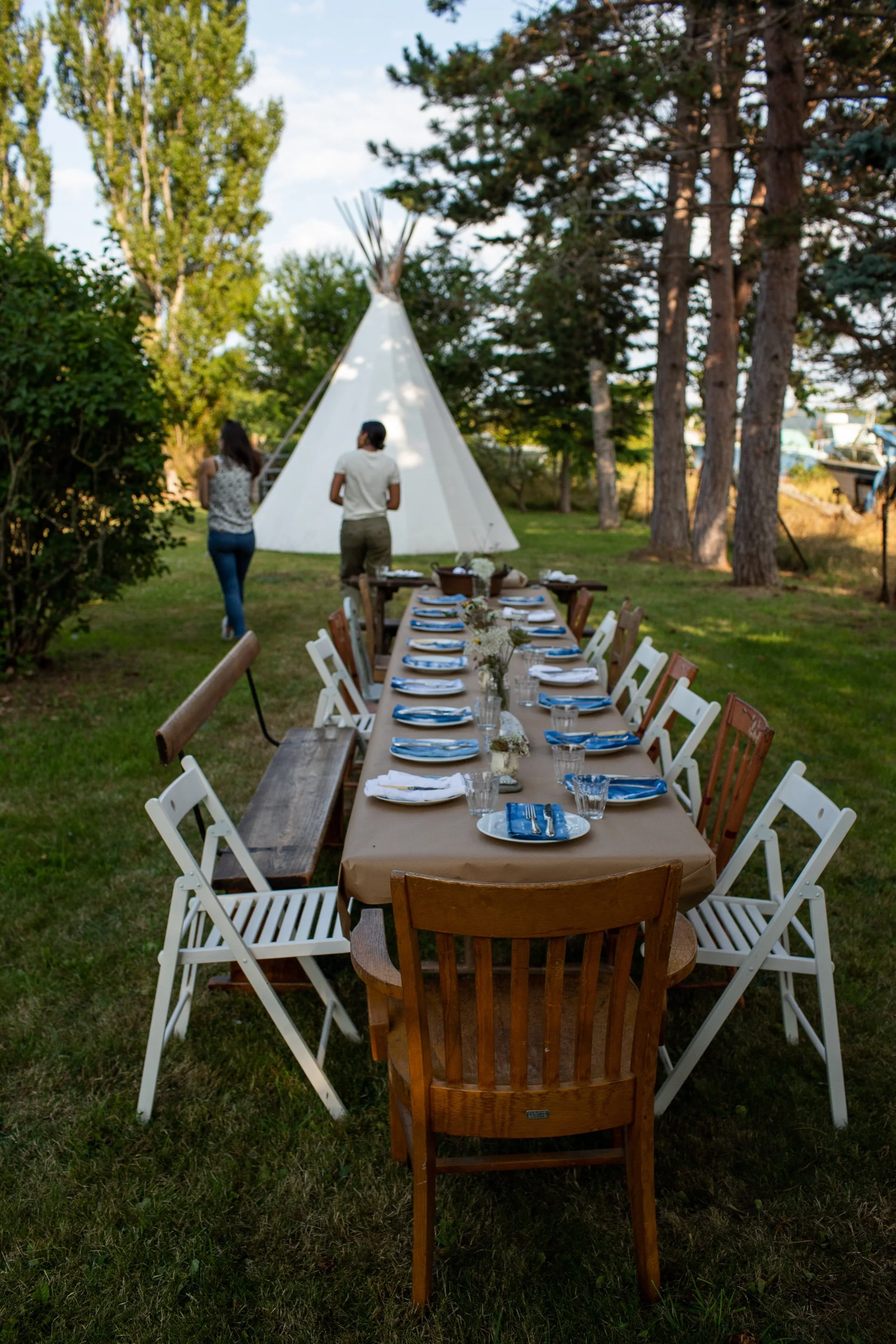 Outdoor gathering with a long dinner table set with plates, glasses, and napkins, with a white teepee and trees in the background.