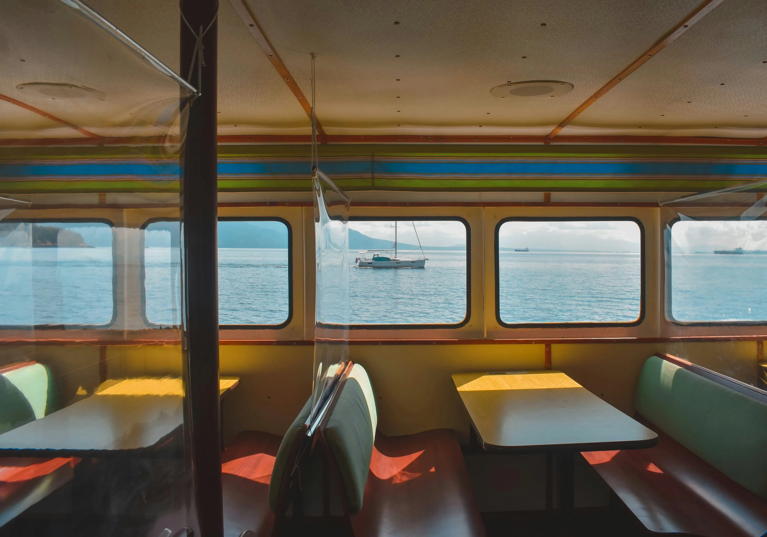 Interior of a boat or ferry with light green seating and wooden tables, viewed through windows showing a body of water with a sailboat and ships in the distance.