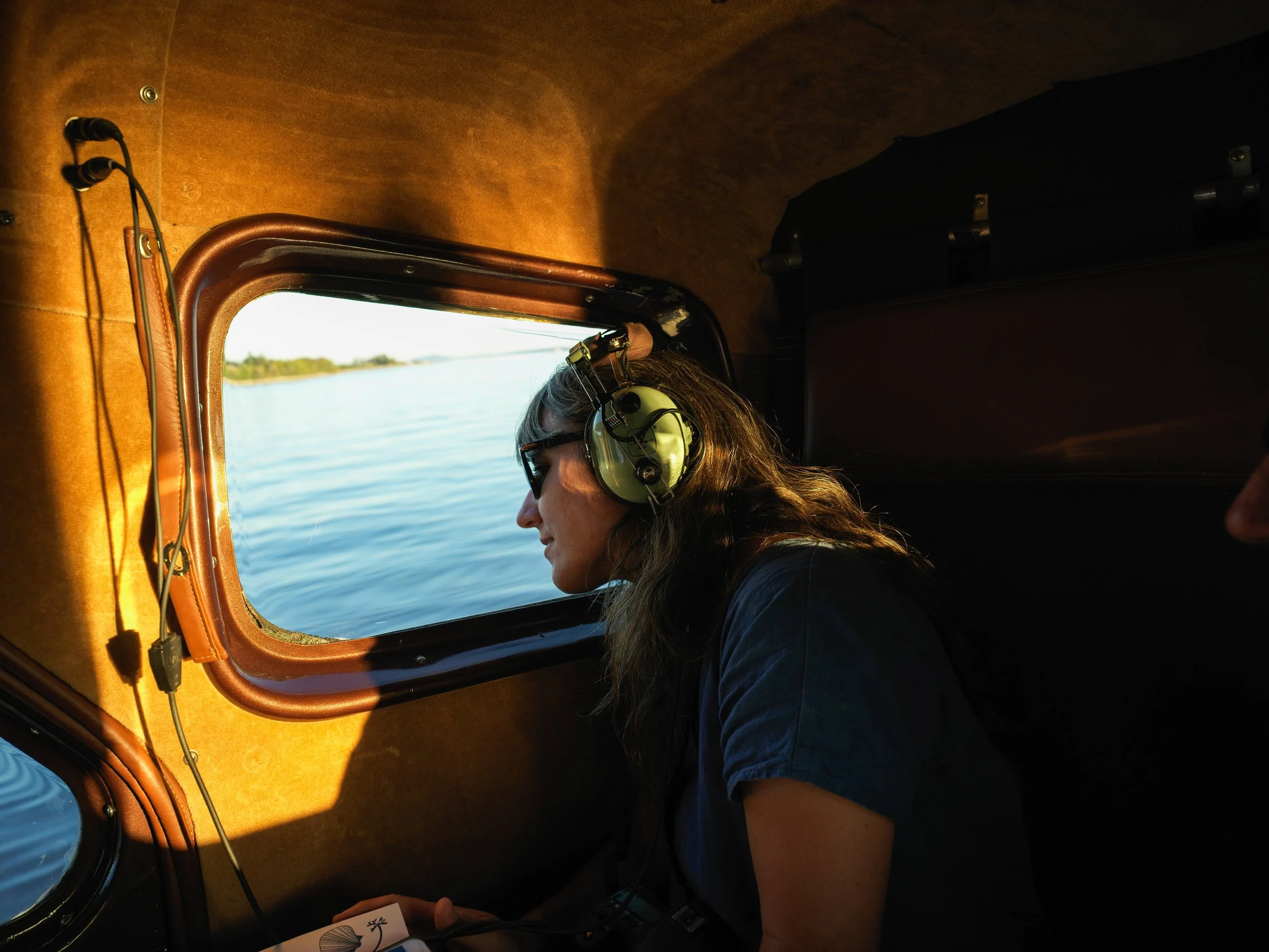A woman wearing headphones and sunglasses looks out of a boat window at a calm body of water with trees in the distance.