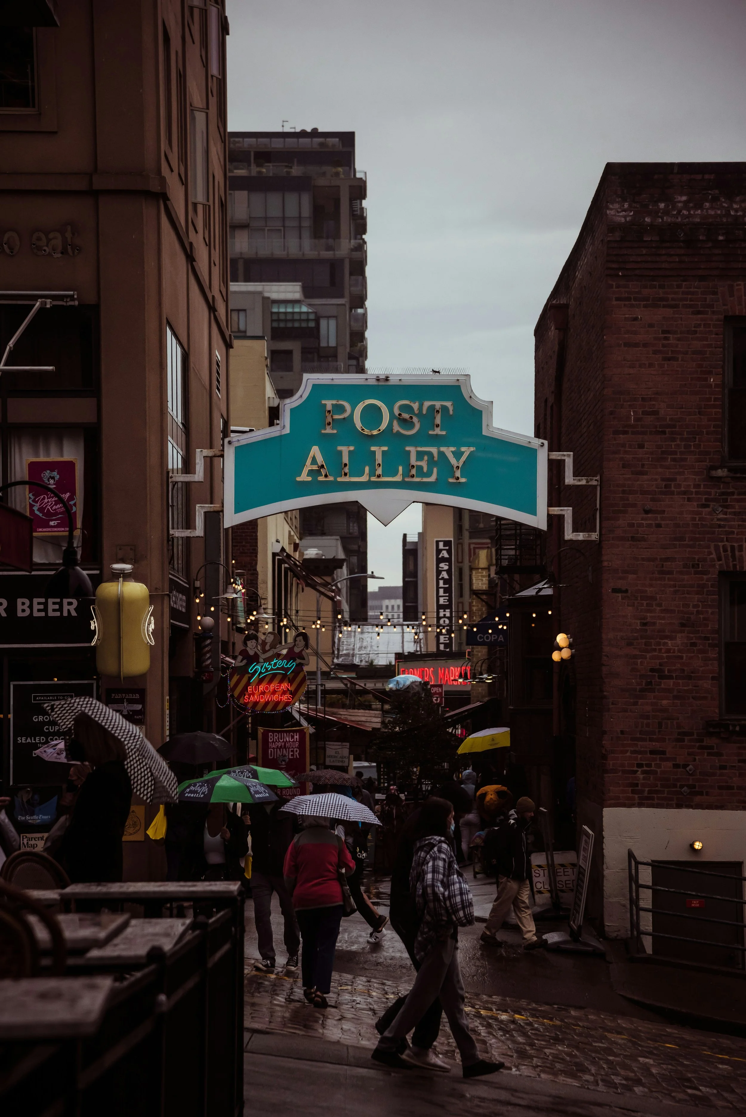 Wet street with people walking under umbrellas in front of Post Alley sign in a city, with buildings and neon signs in the background.