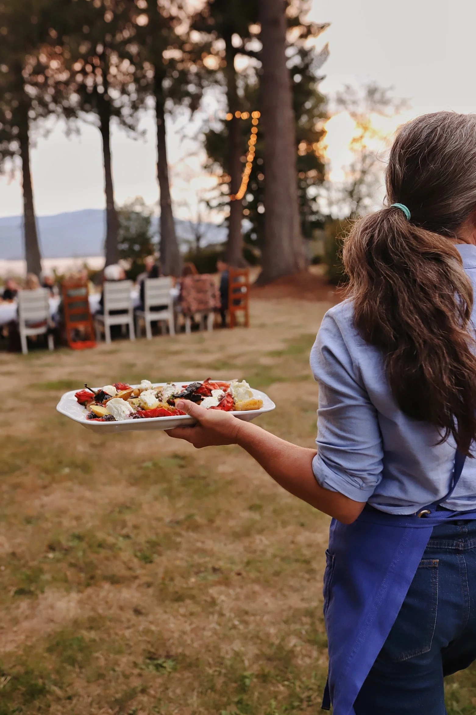 A woman holding a plate of assorted dessert, outdoors at sunset, with a group of people sitting at tables in the background among tall trees.